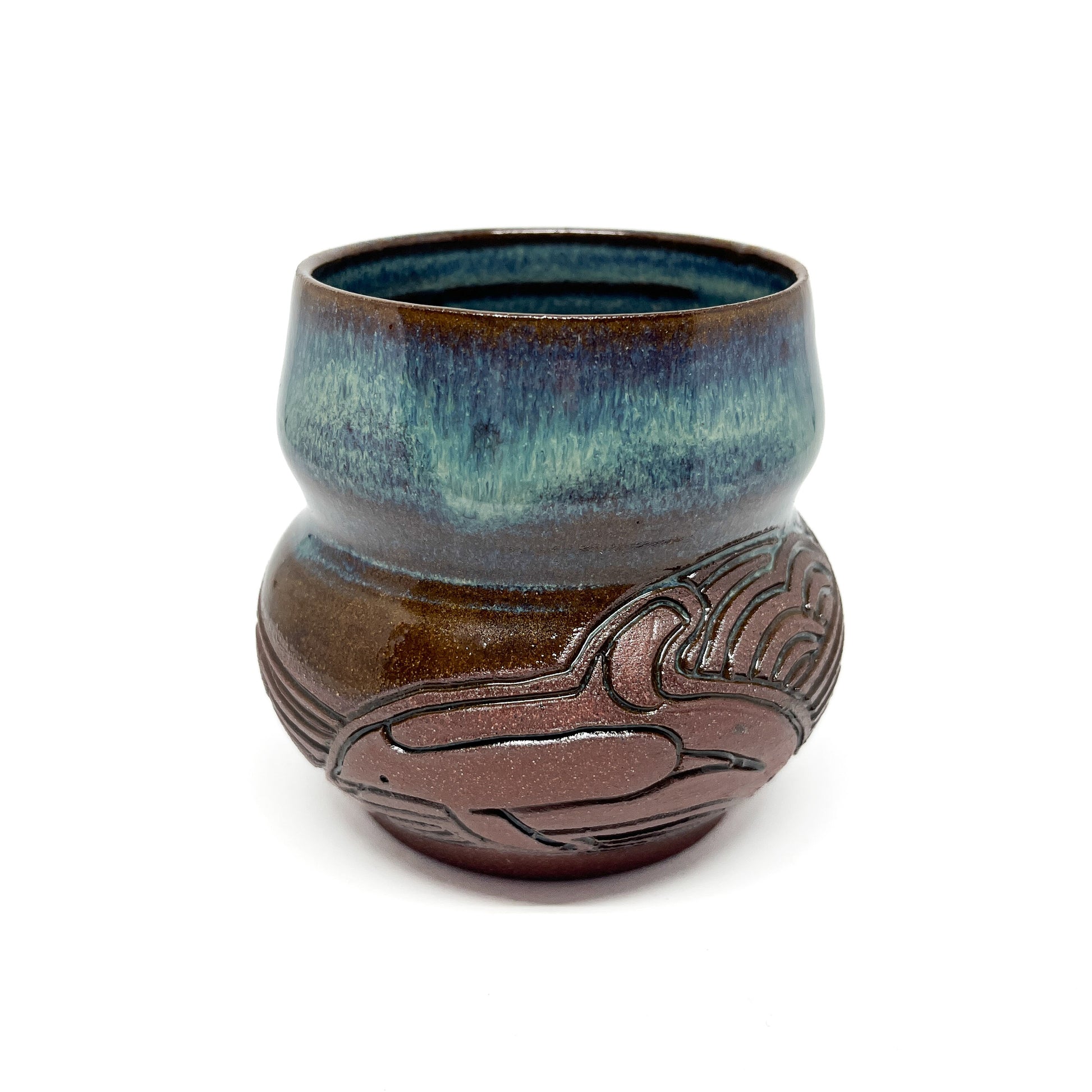 A small, carved ceramic cup with a blue-green top and a brown bottom featuring a stylized whale design.