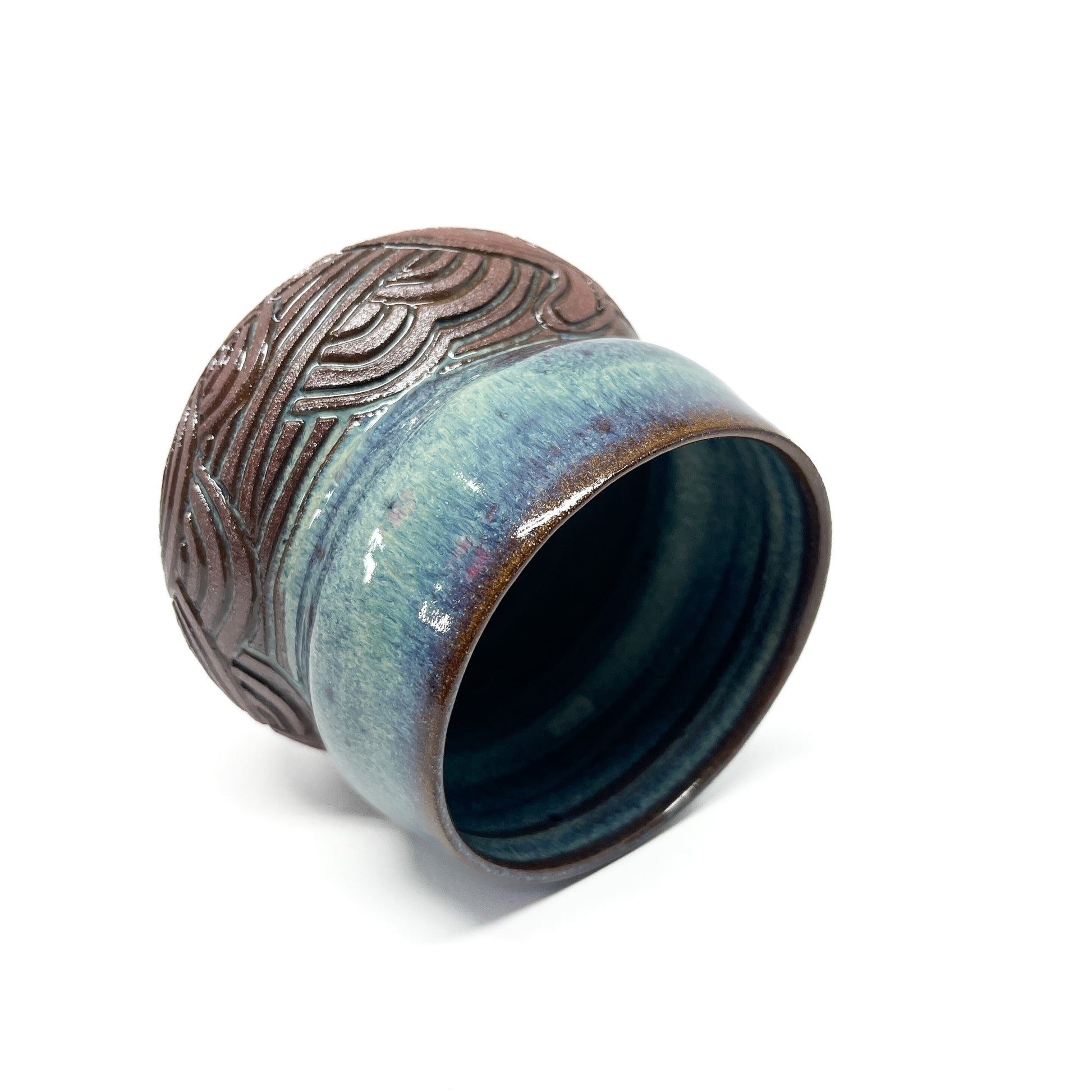 A small, carved ceramic cup with a blue-green top and a brown bottom featuring a textured, abstract design.