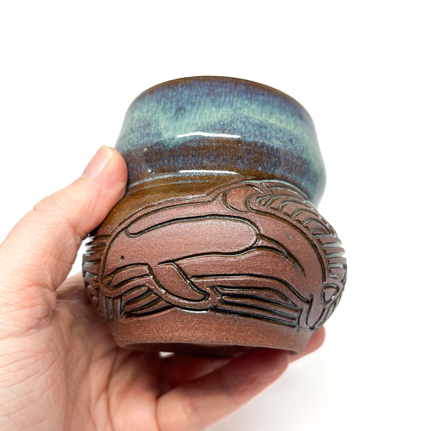 A small, carved ceramic cup with a blue-green top and a brown bottom featuring a carved whale design.