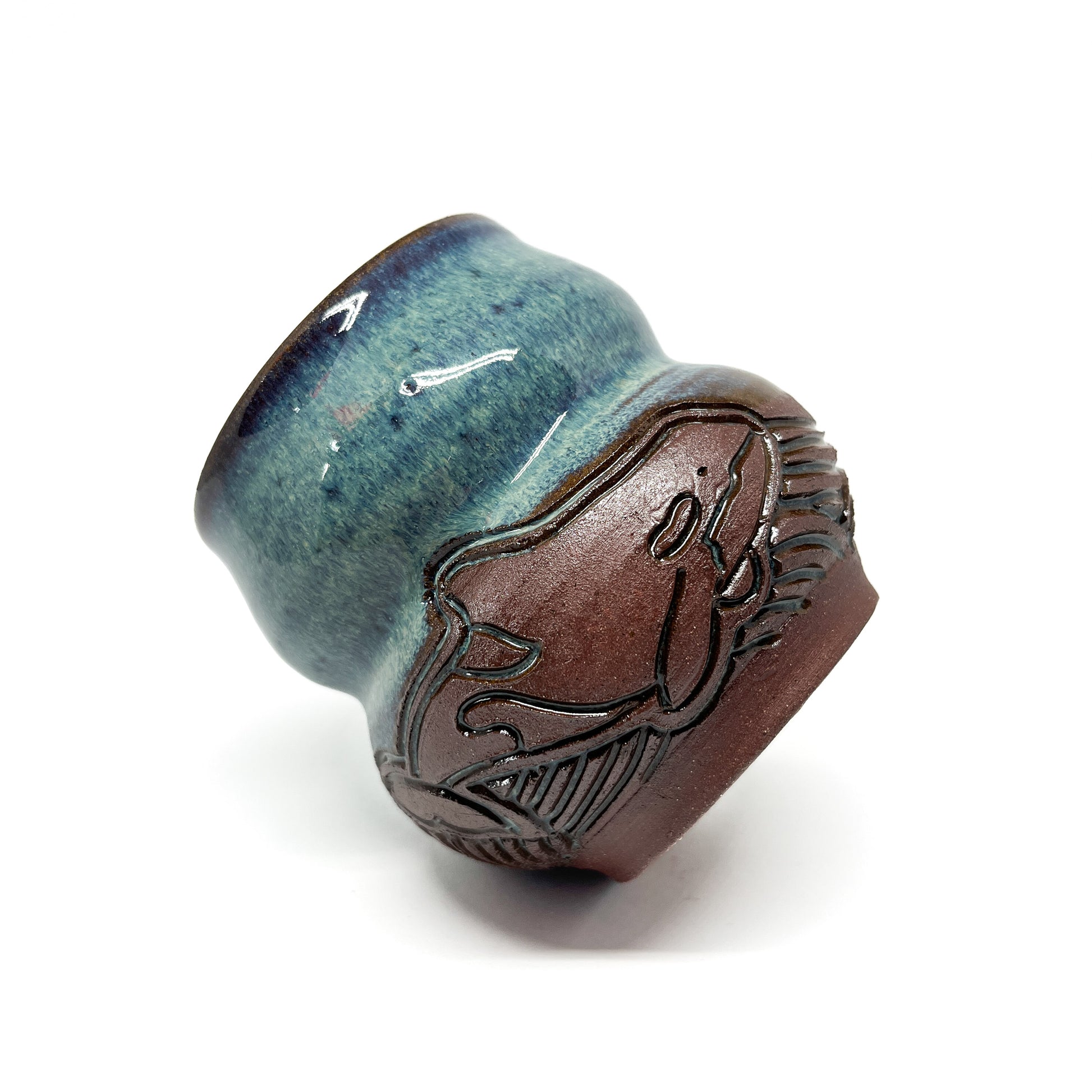 A small, carved ceramic cup with a blue-green top and a brown bottom featuring a carved whale design.