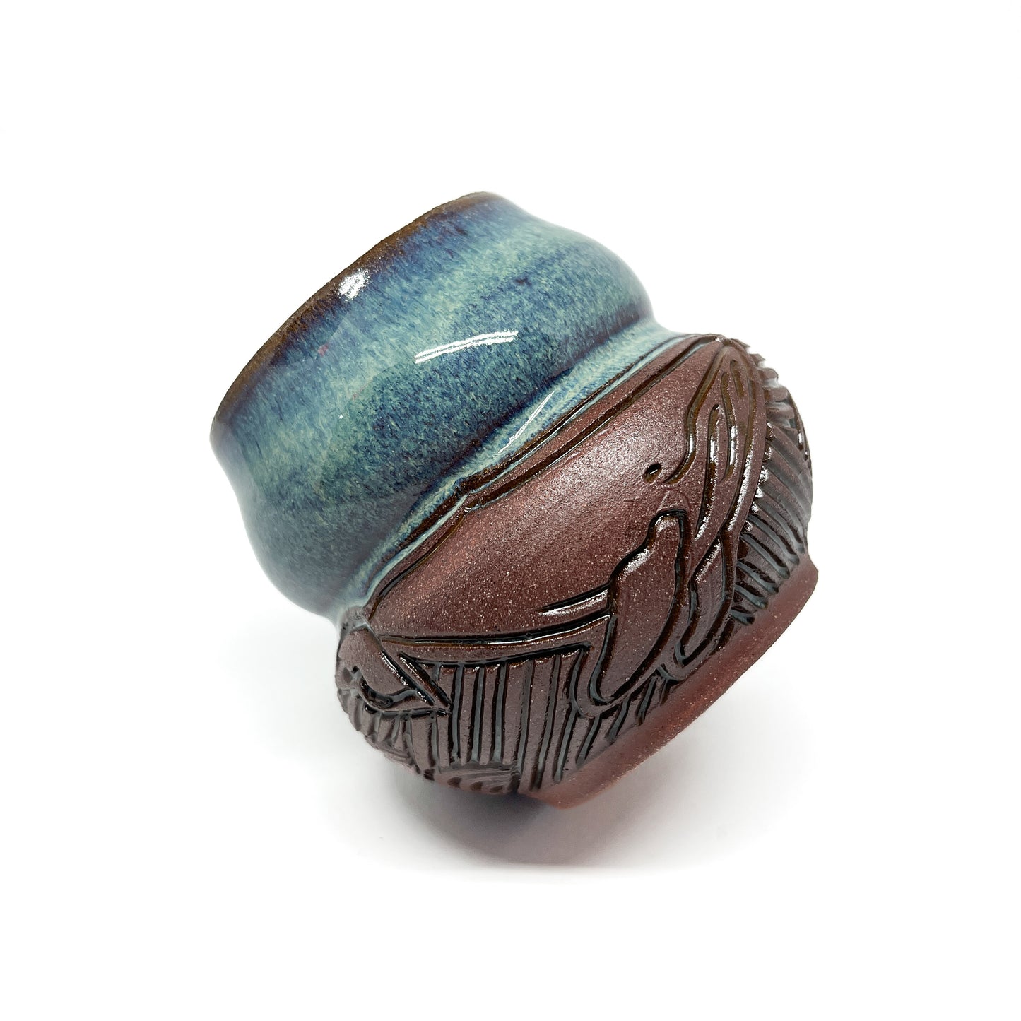 A small, carved ceramic cup with a blue-green top and a brown bottom featuring a carved whale design.