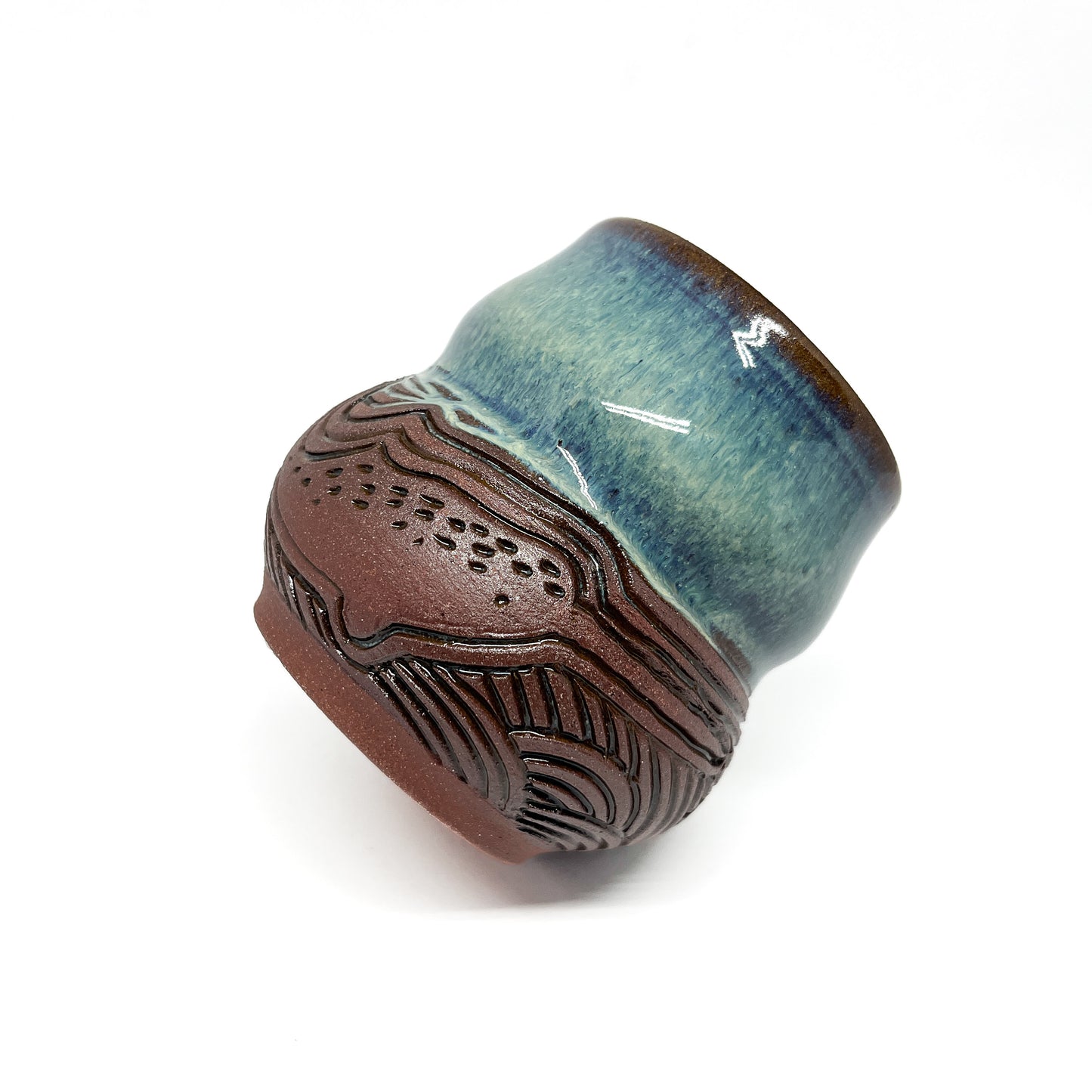 A small, carved ceramic cup with a blue-green top and a brown bottom featuring a wave design.