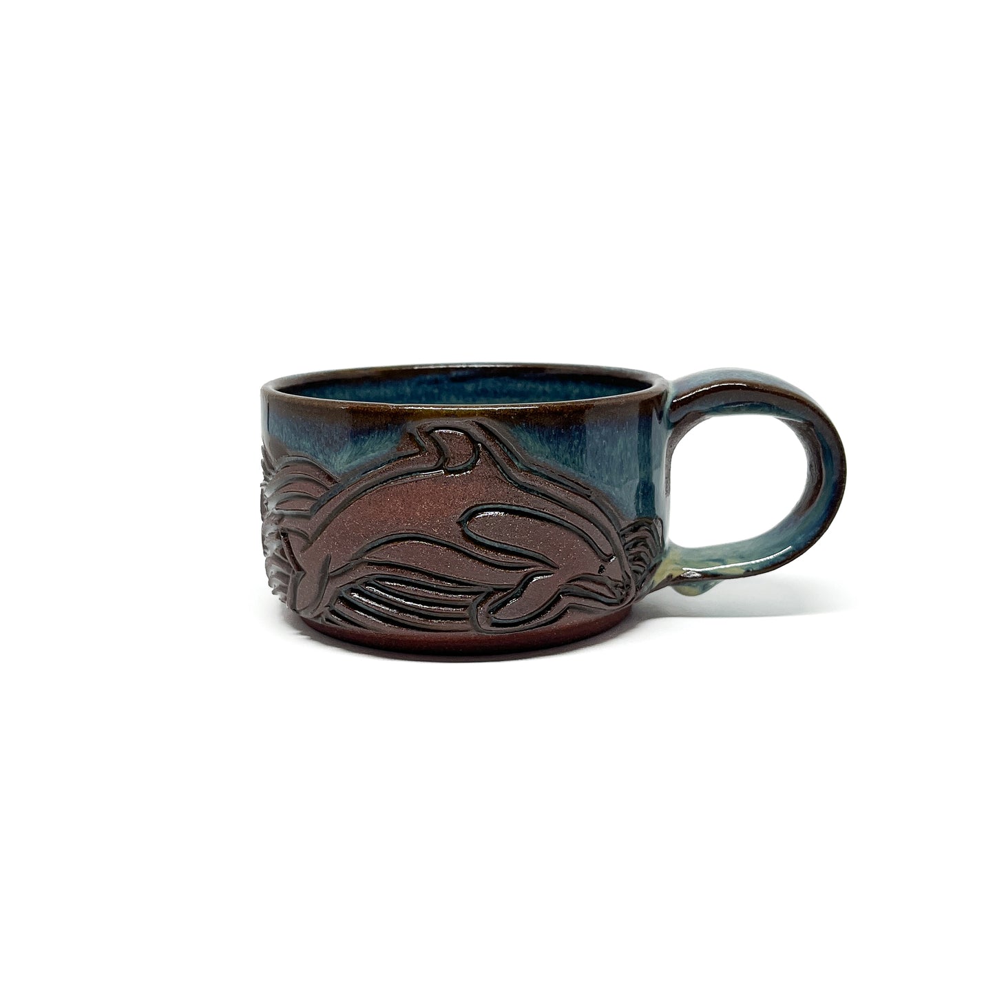 Ceramic mug with a carved whale and wave design in brown against a glossy blue-green glaze and a rounded handle.
