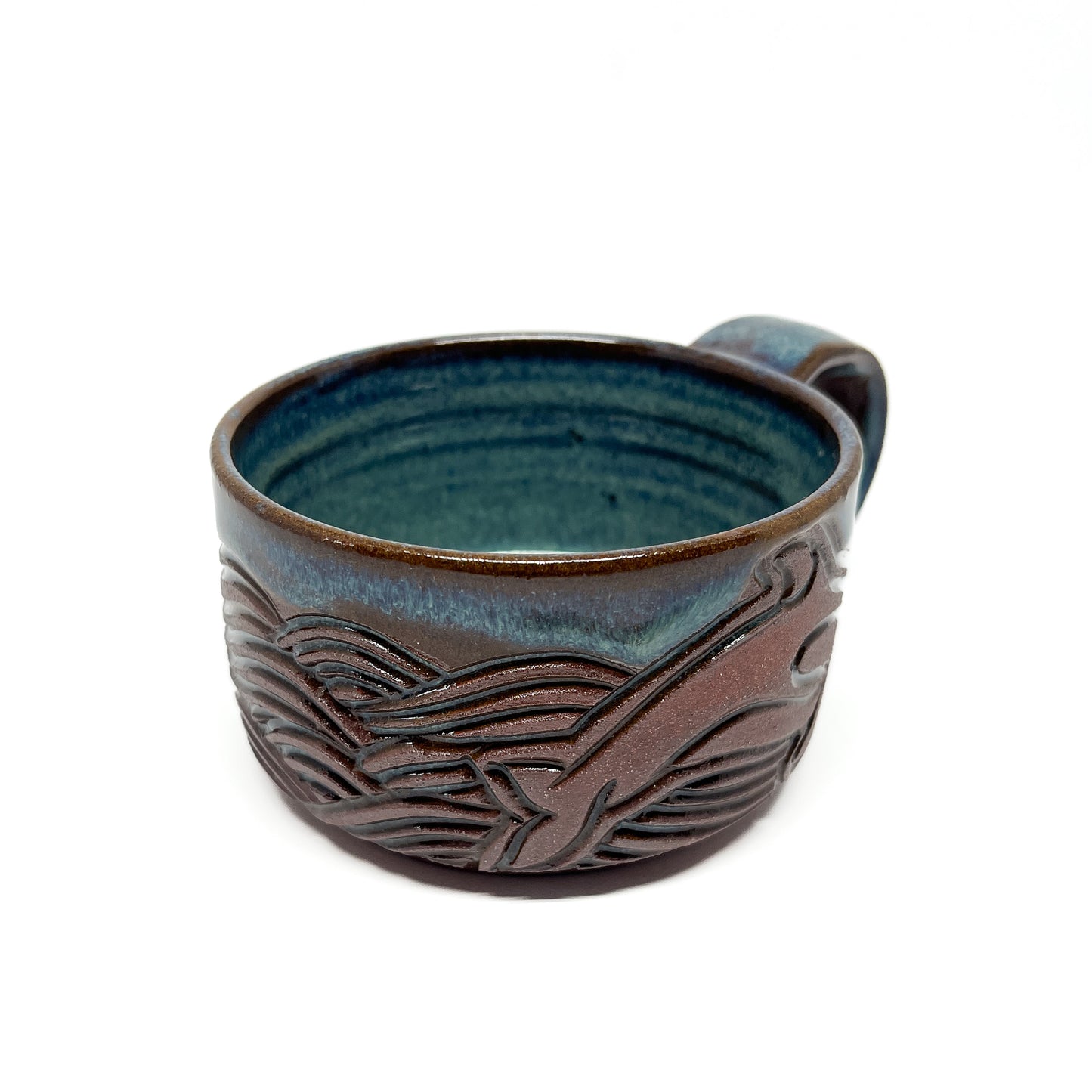 Ceramic mug with a carved whale and wave design in brown against a glossy blue-green glaze and a rounded handle.