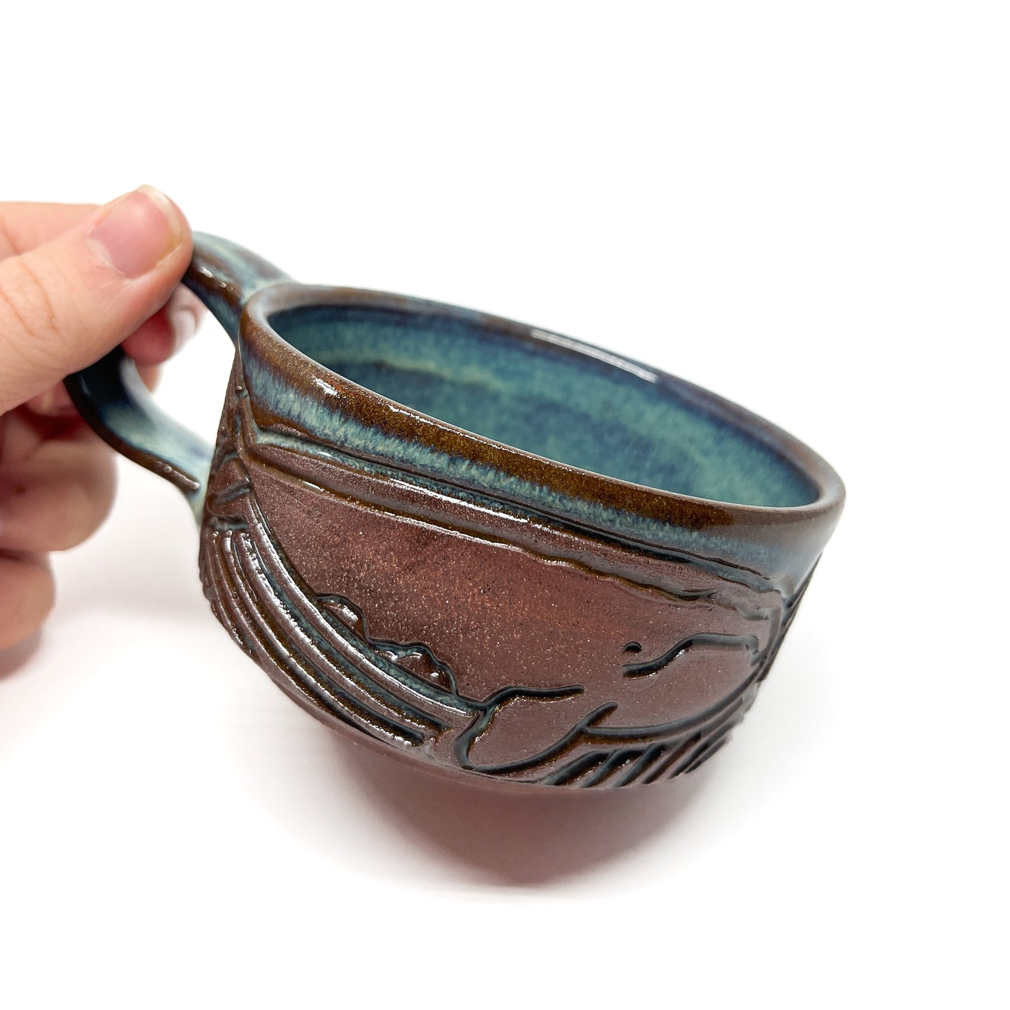 Person holding a handmade ceramic cup with engraved whale design and a glazed interior and rim.