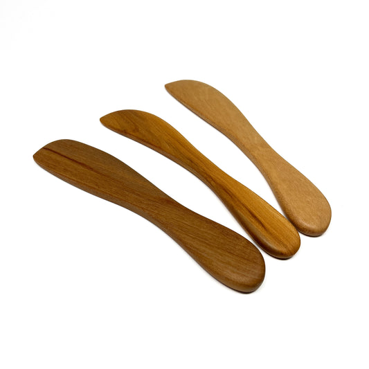 Three wooden spread knives in varying shades, angled on a white surface.