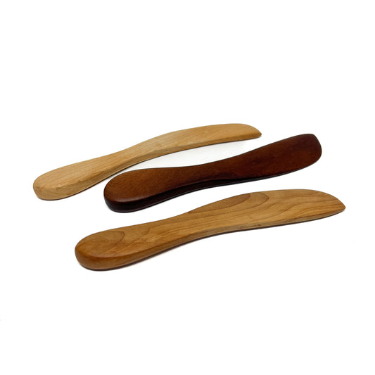 Three wooden spread knives in varying shades, angled on a white surface.