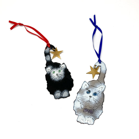 Two cat Christmas ornaments, one black and white, one gray and white, each holding a gold star by its tail with a ribbon for hanging.