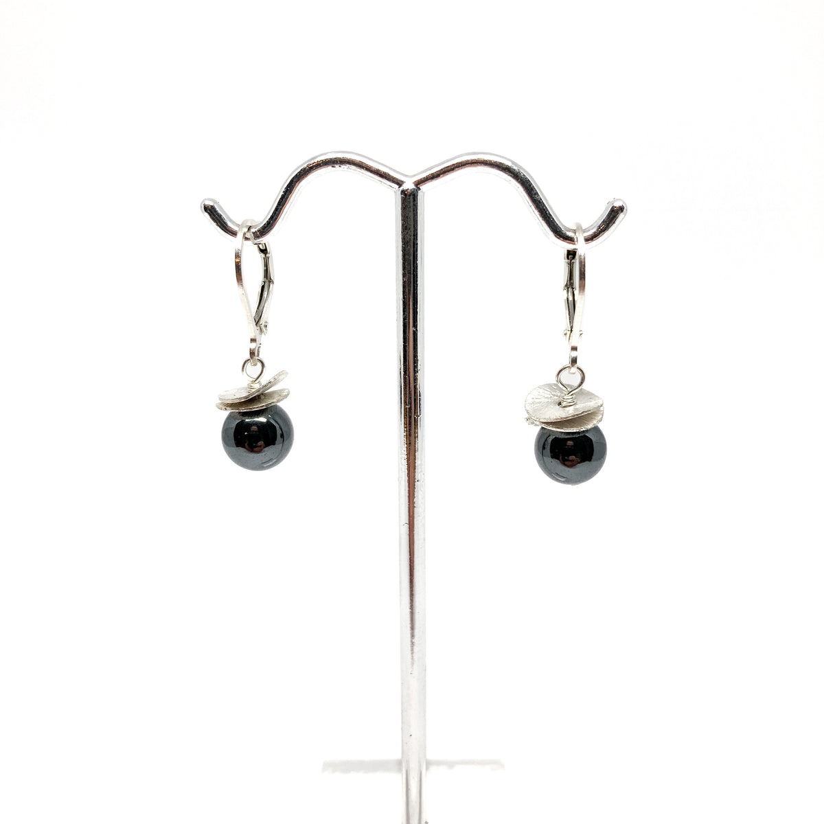 A pair of silver lever-back earrings with dark beads and a textured, petal-like design, displayed on a metal earring stand. The earrings have a secure lever closure.