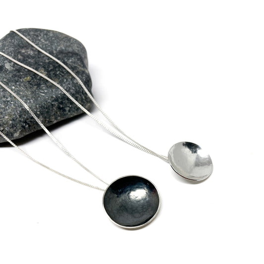 Two Sister Moons pendants, one oxidized dark, one shiny silver, rest on a gray stone.