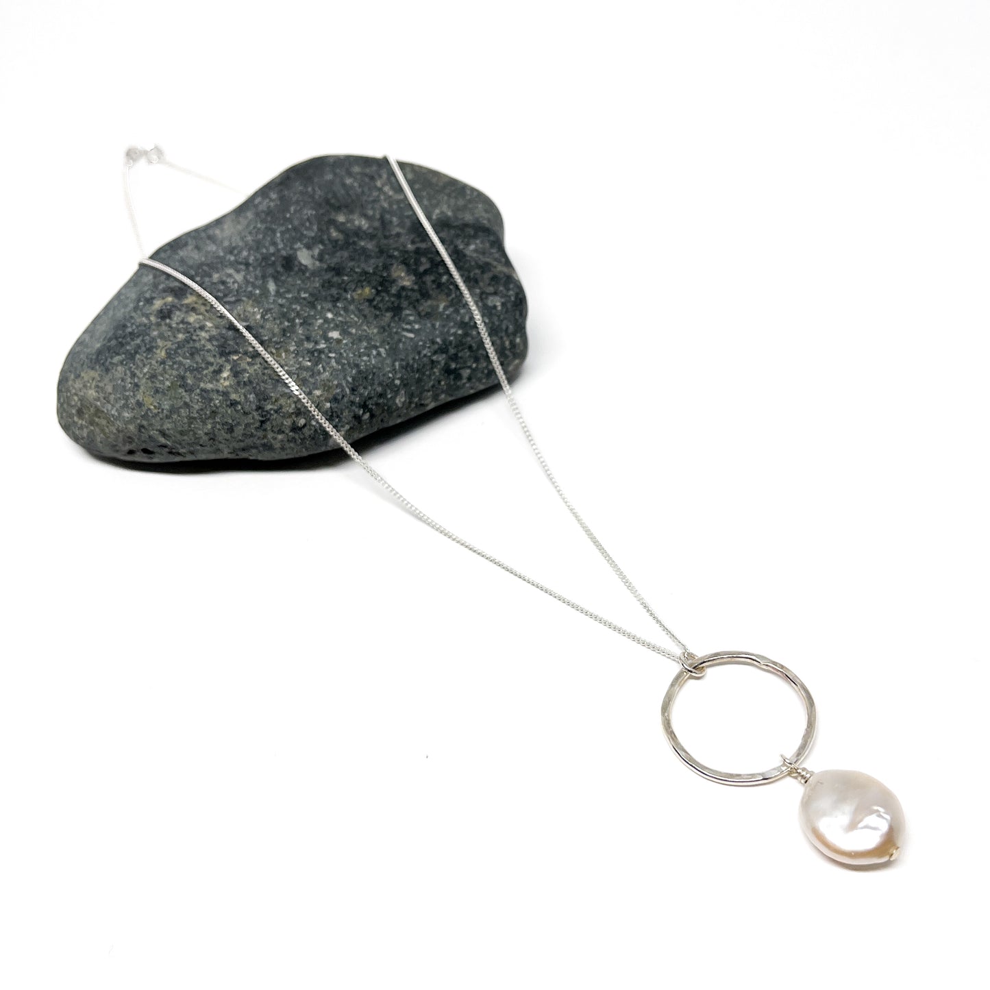 Silver loop necklace with a single baroque pearl pendant resting on a textured stone.