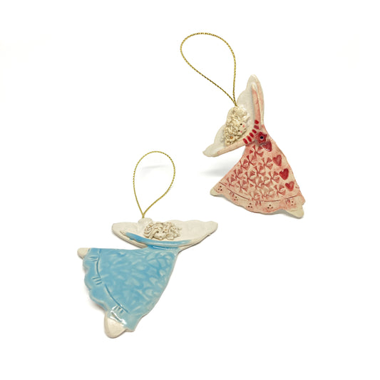 Two angel ornaments with gold strings, one pink with hearts and one blue, viewed from above.