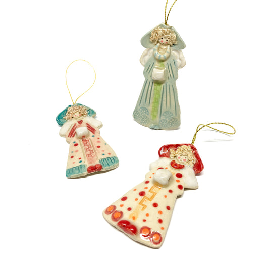 Three angel ornaments with hoods and gold strings, each with a unique design and color.