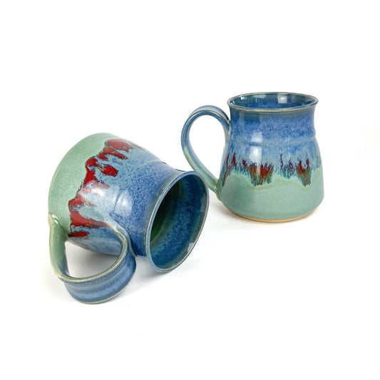 Two ceramic mugs with a blue-to-green gradient and red accents, one tilted on its side.