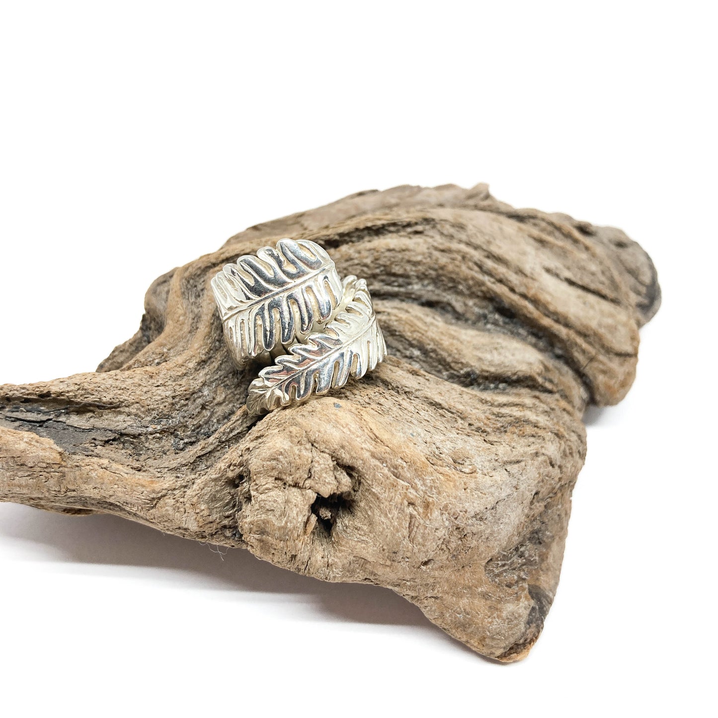 Silver ring shaped like a stylized fern leaf, resting on a textured piece of driftwood.