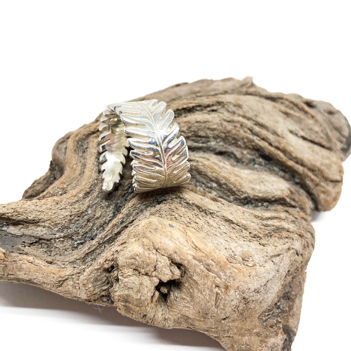 Silver cuff bracelet shaped like stylized fern leaves, resting on a piece of driftwood.