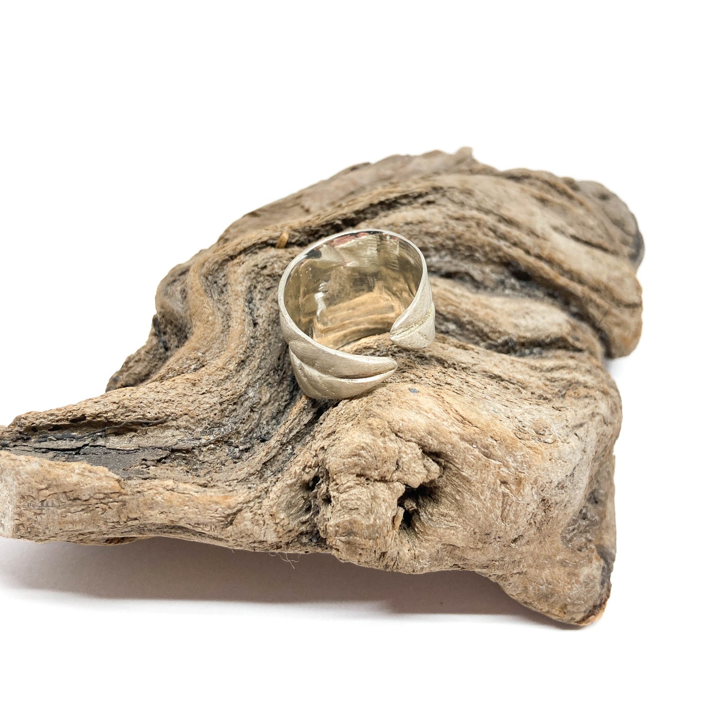 Silver ring with a textured, overlapping leaf design, resting on a piece of textured driftwood.