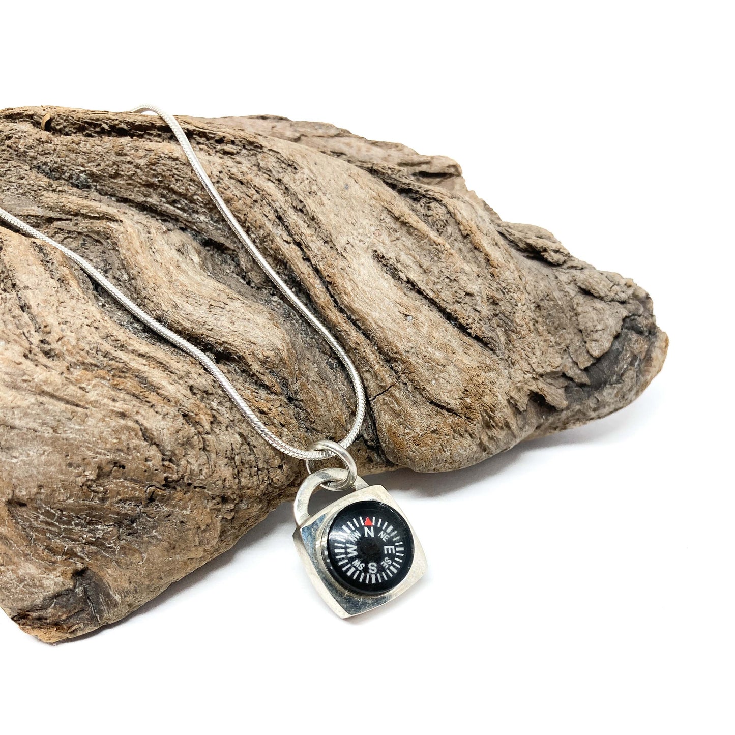 A silver necklace with a compass pendant rests on a textured piece of driftwood. The compass face shows cardinal directions.