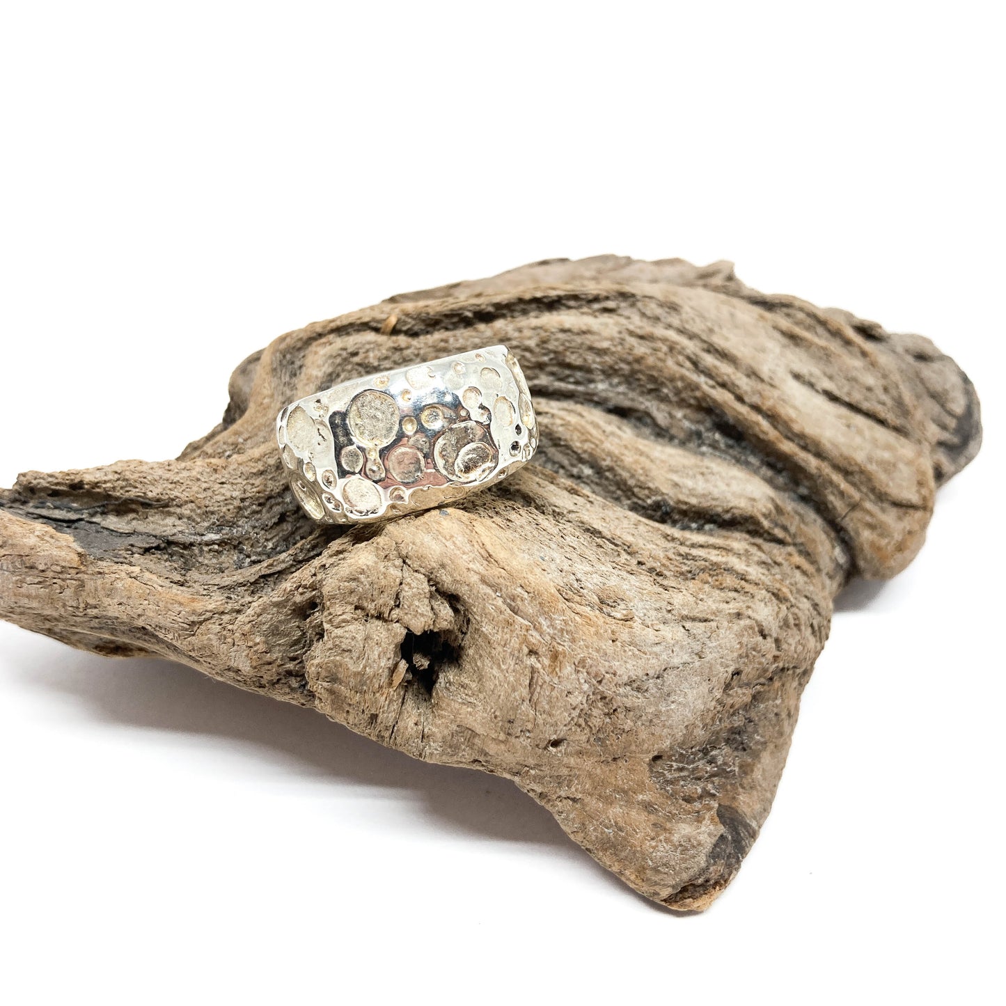 A wide sterling silver ring with a rustic, textured surface rests on a piece of weathered driftwood.
