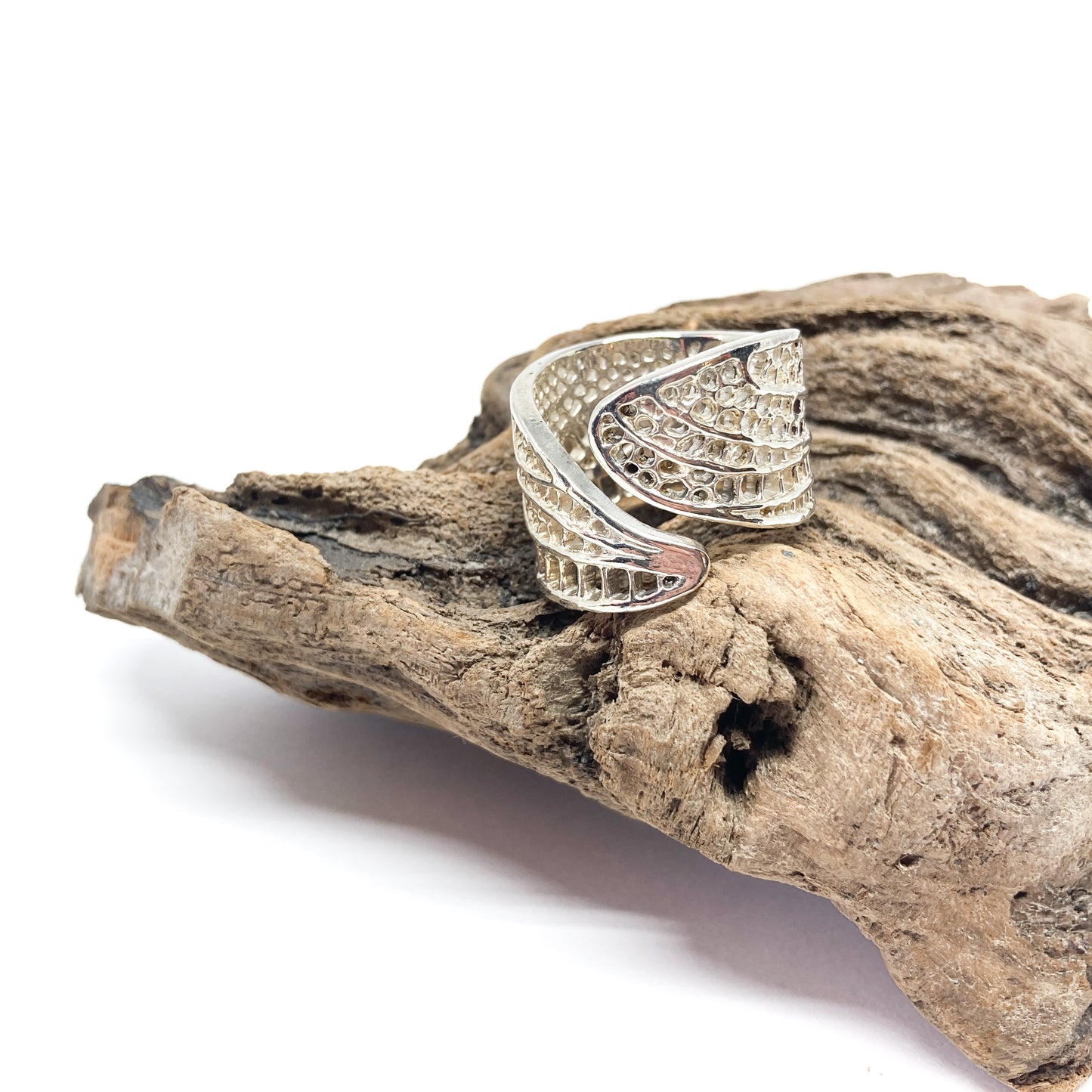 A shiny silver ring shaped like detailed dragonfly wings rests on a textured piece of driftwood.