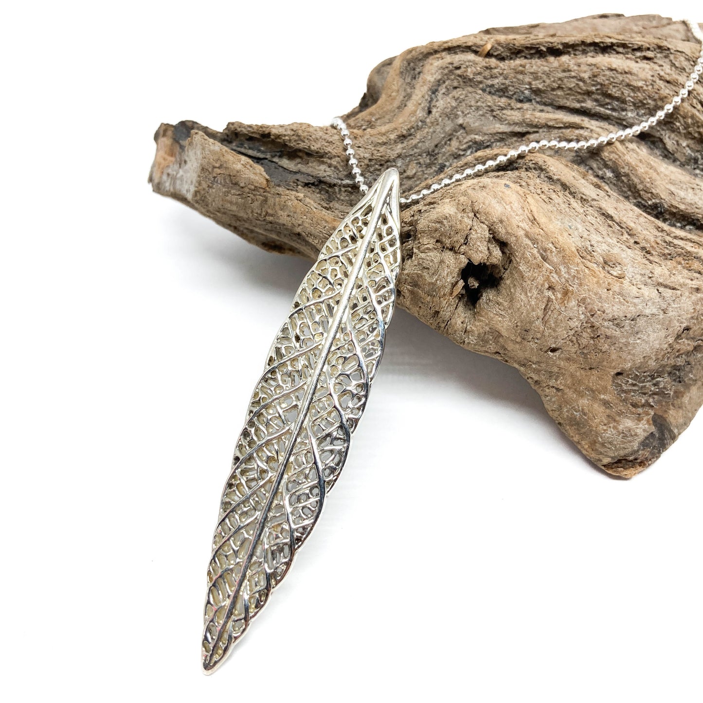 A sterling silver necklace with a detailed, openwork leaf pendant rests on a piece of textured driftwood.