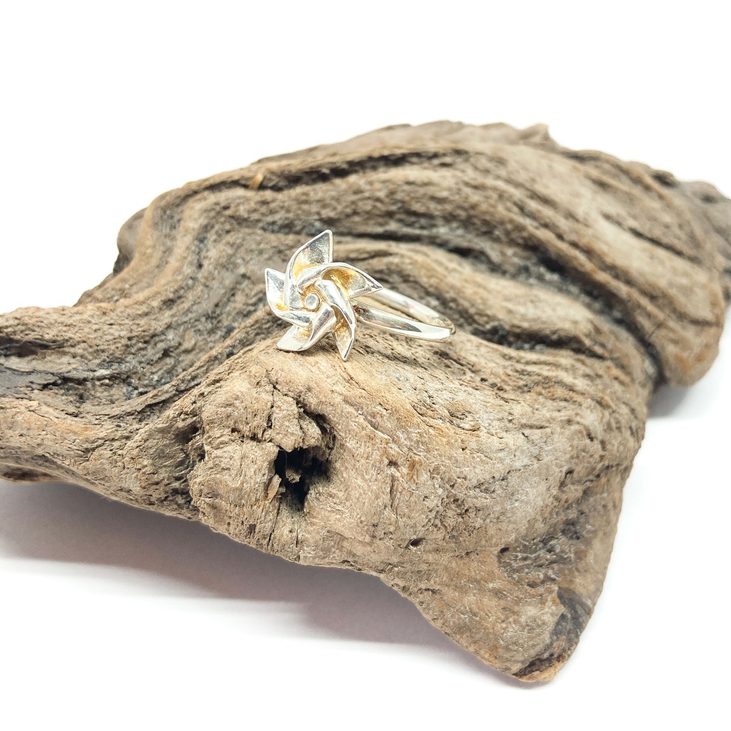 A shiny silver ring with a three-dimensional, stylized floral or pinwheel design rests on a piece of textured driftwood.
