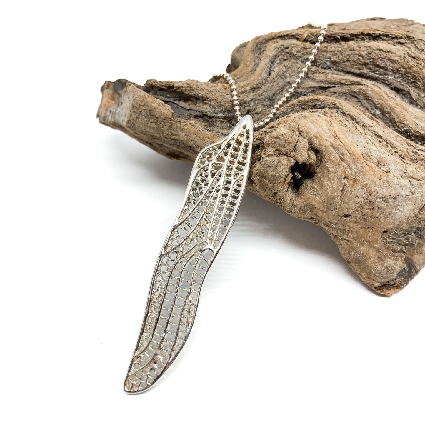 A silver necklace with a detailed dragonfly wing pendant rests diagonally on a textured piece of driftwood.