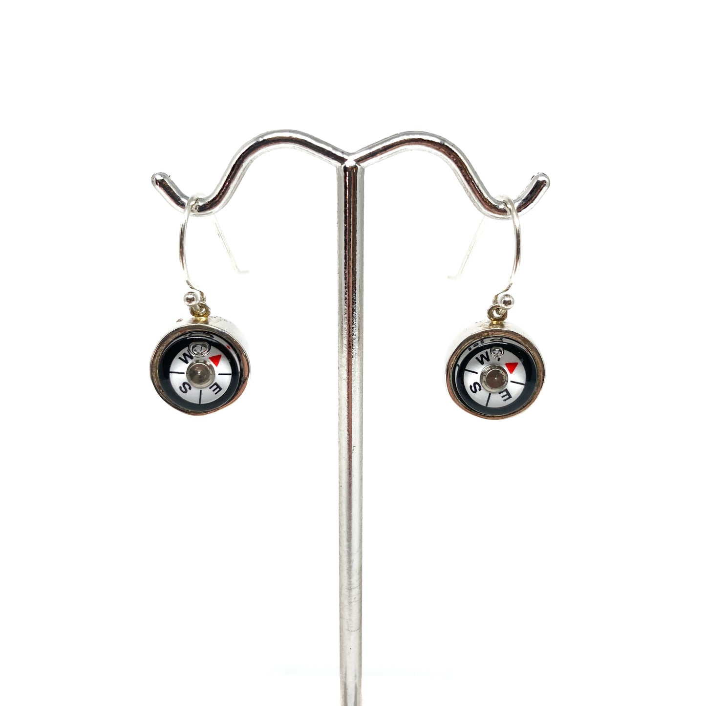 Two shiny silver dangle earrings featuring mini working compasses with cardinal directions hang from a metal stand.