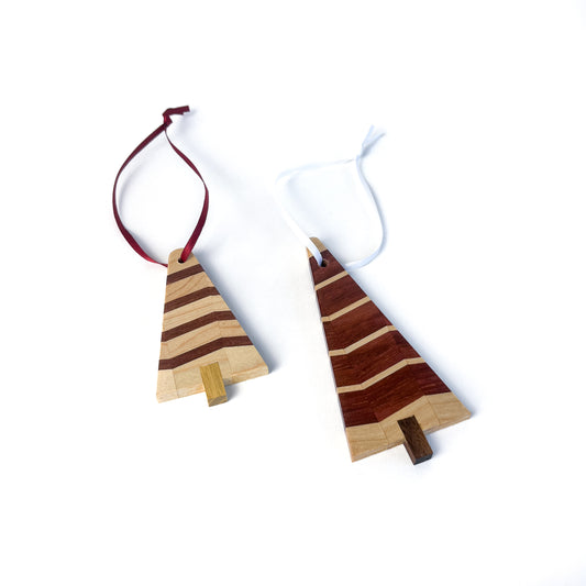 A pair of wooden Christmas tree ornaments with light and dark wood stripes and contrasting trunks, one with a red ribbon and one with a white ribbon.