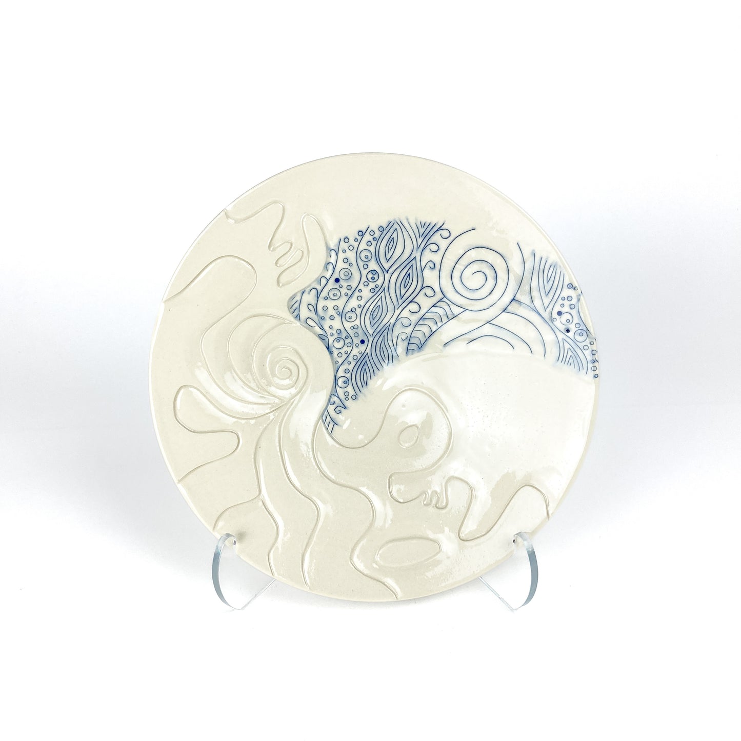 Wide handmade pottery plate on a clear stand, featuring carved white abstract shapes contrasting with blue swirling line art.