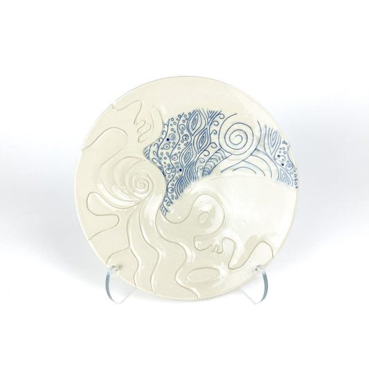Wide handmade pottery plate on a clear stand, featuring carved white abstract shapes contrasting with blue swirling line art.