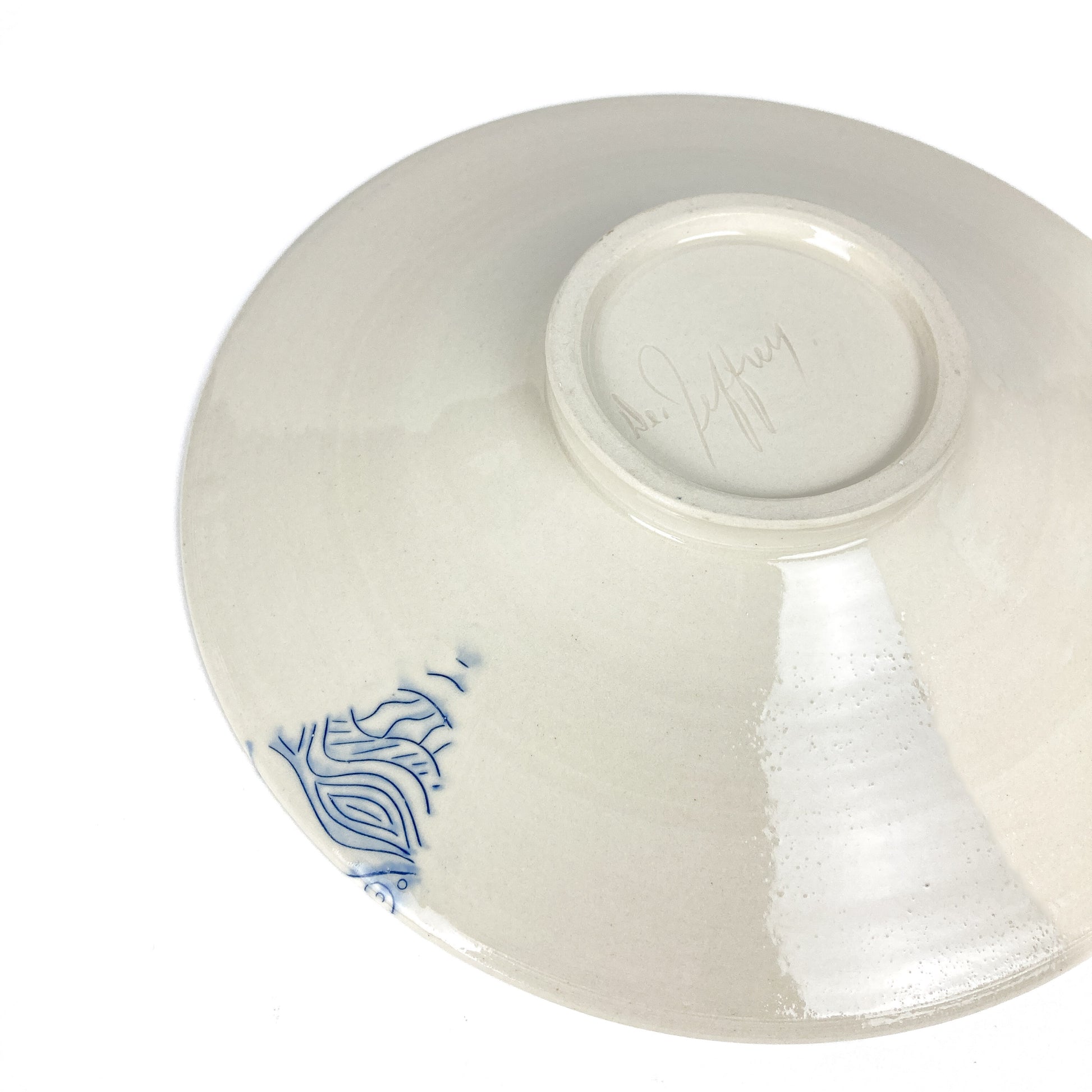 Underside of a wide ceramic plate with a small blue abstract design and a signature.