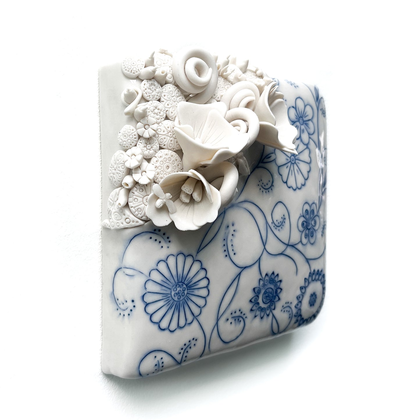 Angled view of a square ceramic tile with blue floral line art and raised, textured white flowers.