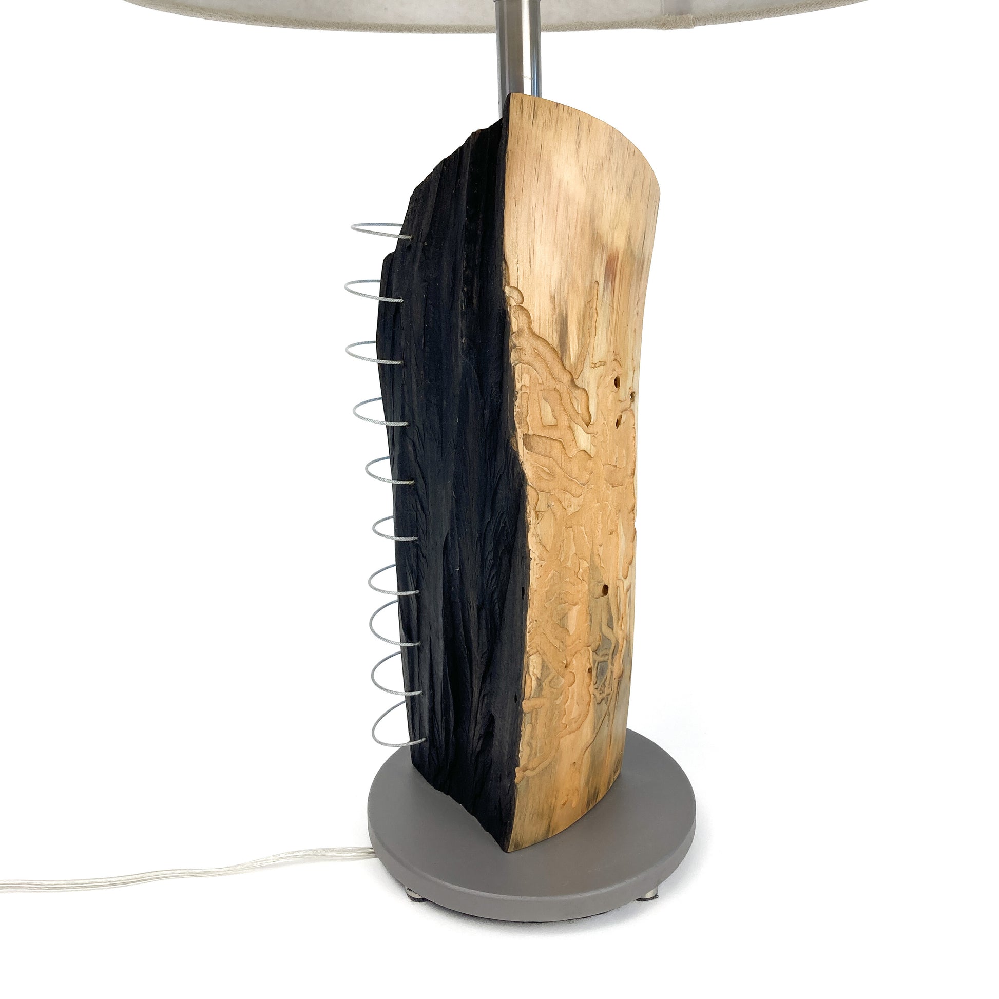 Close-up of a lamp base made of natural wood with a charred blackened section and a metal spiral detail, mounted on a round stand.