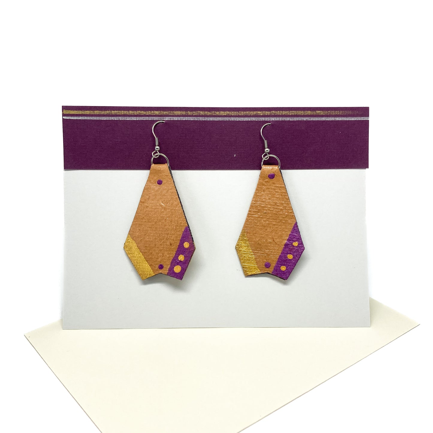 Two tan geometric earrings with gold and purple accents hang from silver hooks on a display card.