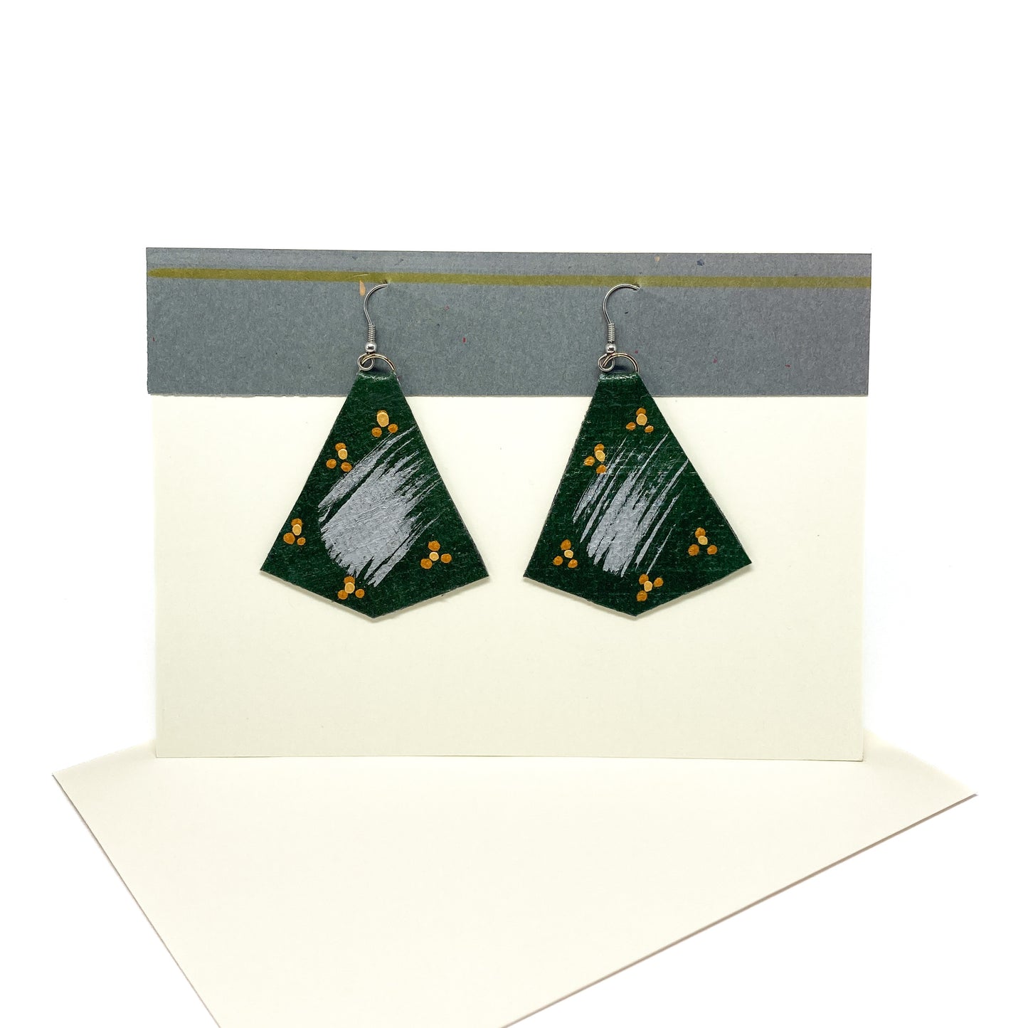 Two dark green, gold, and silver geometric earrings hang from silver hooks on a display card. They feature abstract brushstrokes.