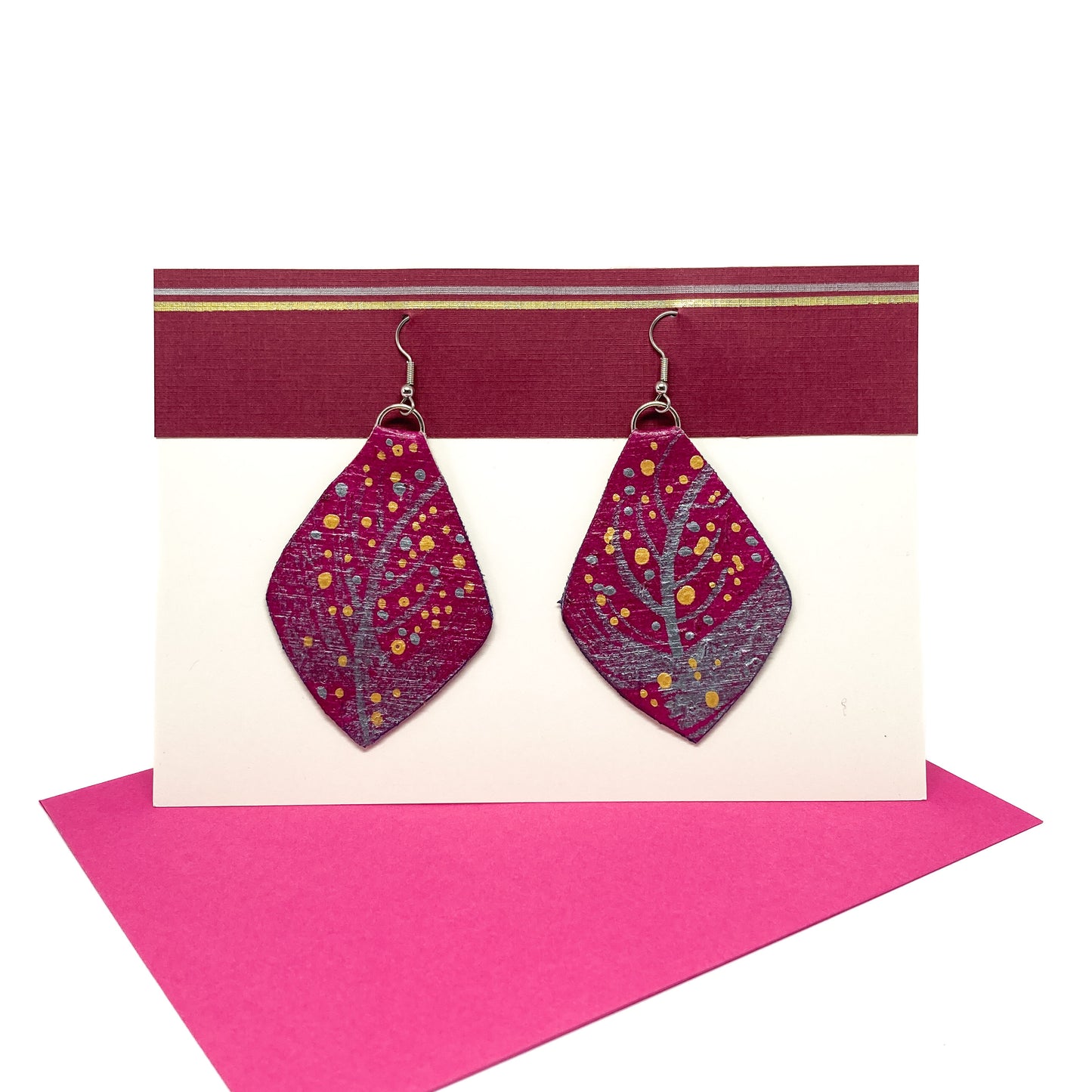 Two pink geometric earrings with silver branch-like designs and gold dots hang from silver hooks on a display card.