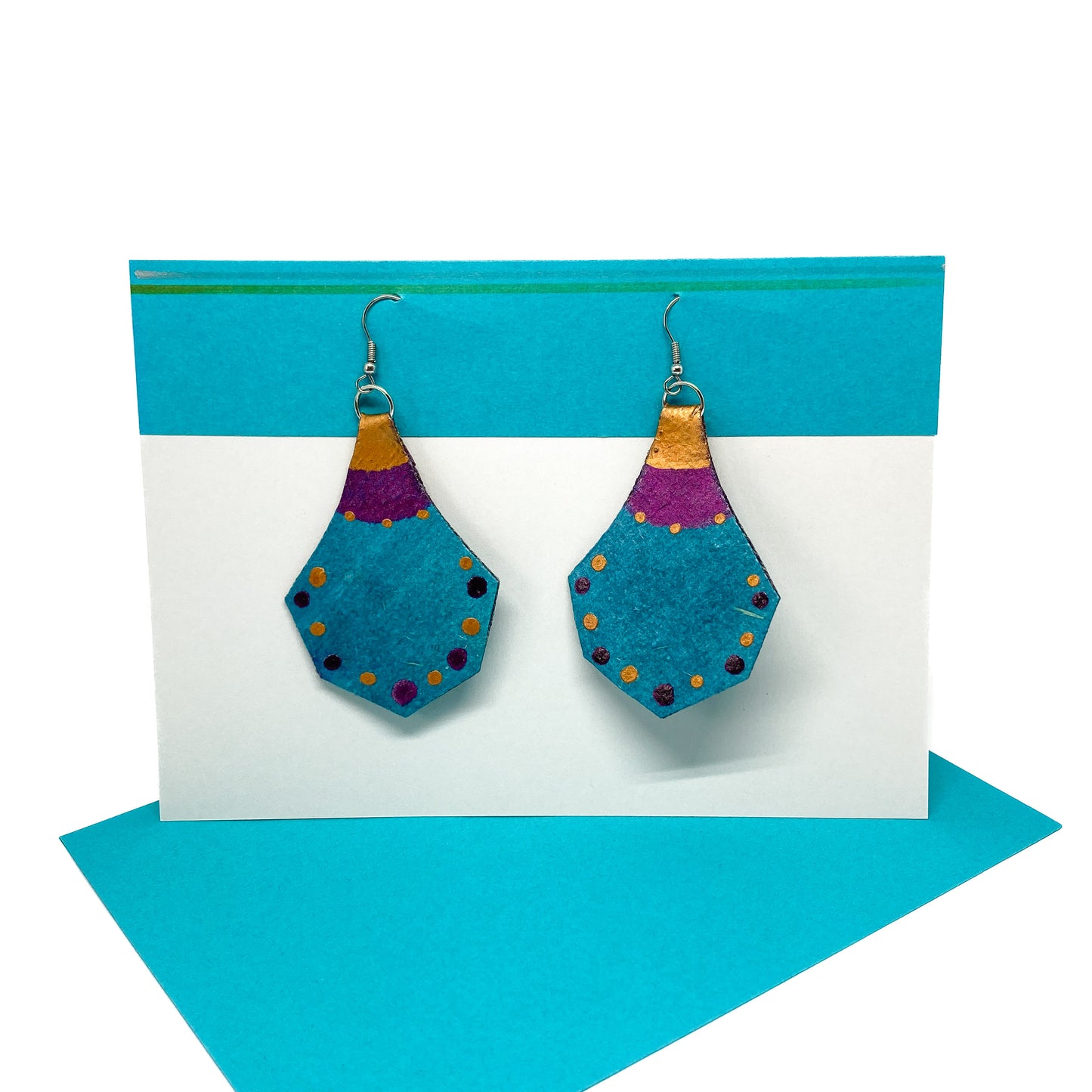 Two teal, purple, and gold geometric earrings with a unique shape and dotted patterns hang from silver hooks on a display card.