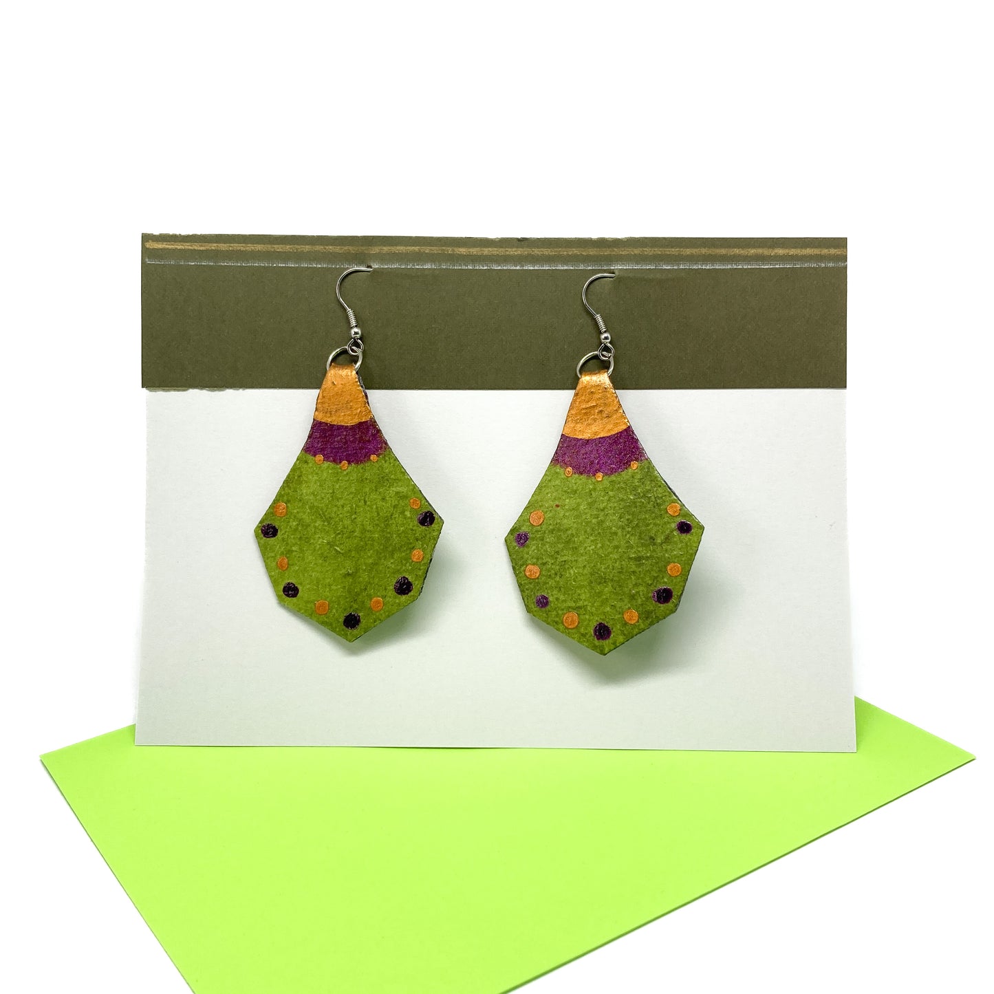 Two green, purple, and gold geometric earrings with a unique shape and dotted patterns hang from silver hooks on a display card.