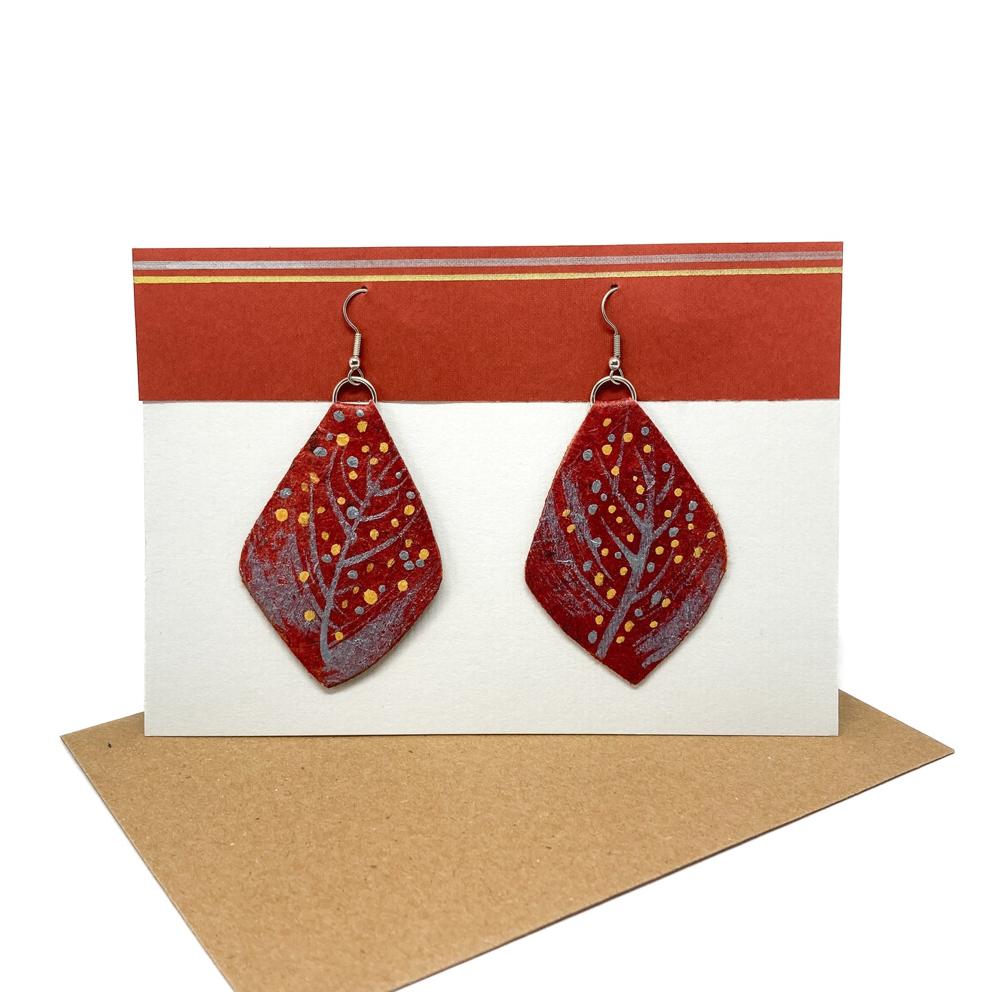 Two red geometric earrings with silver branch-like designs and gold dots hang from silver hooks on a display card.