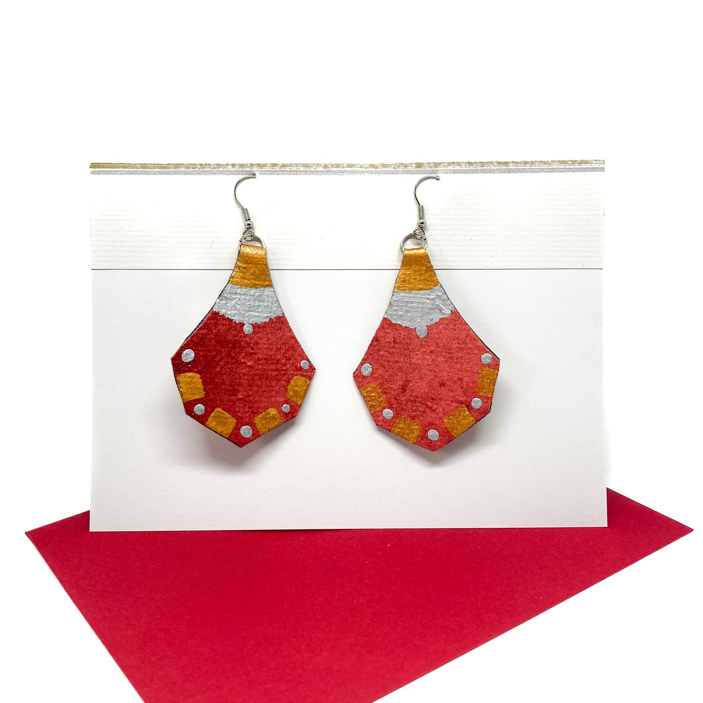 Two red, gold, and silver geometric earrings with a unique hexagonal shape hang from silver hooks on a display card.