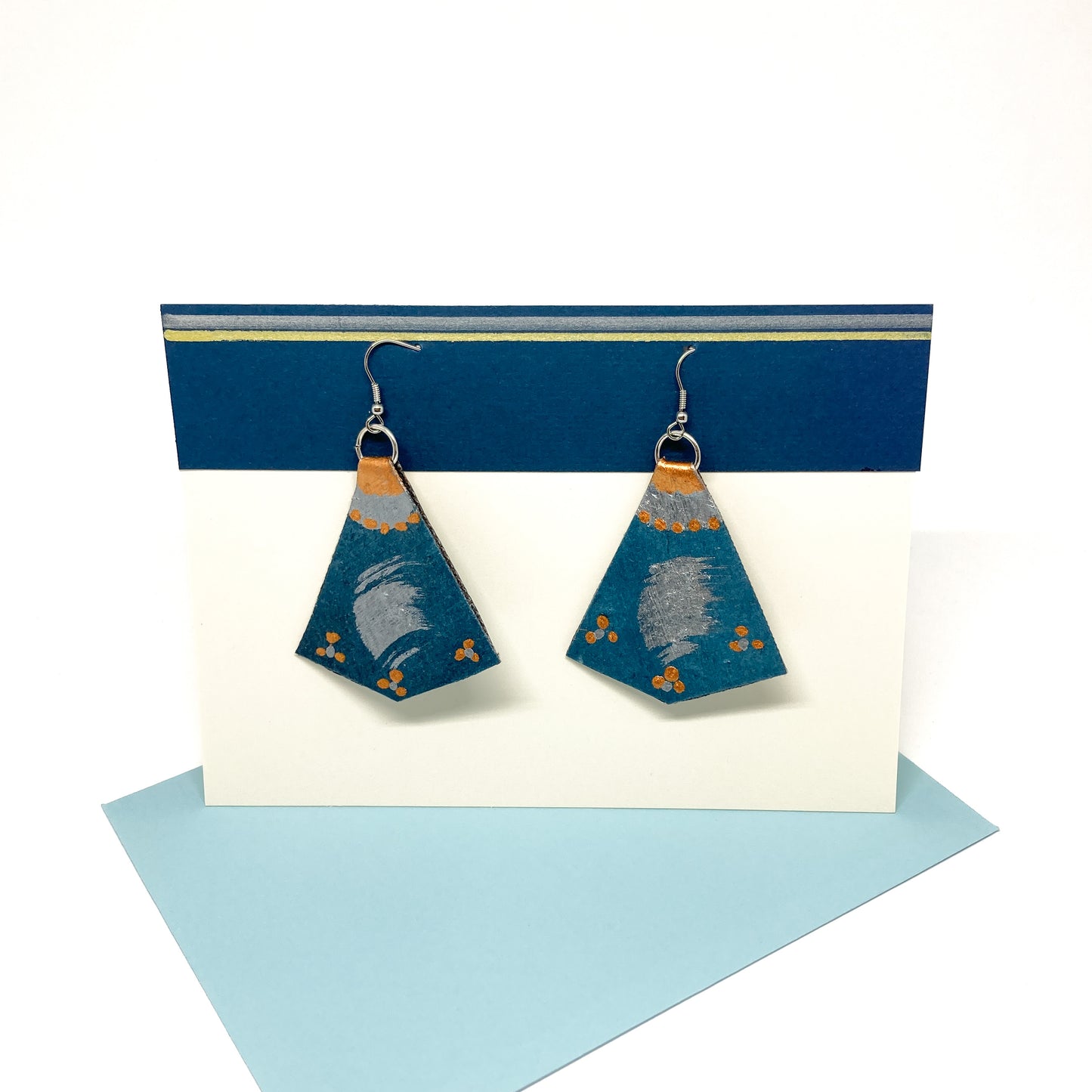 Two teal, copper, and silver geometric earrings with abstract designs hang from silver hooks on a display card.