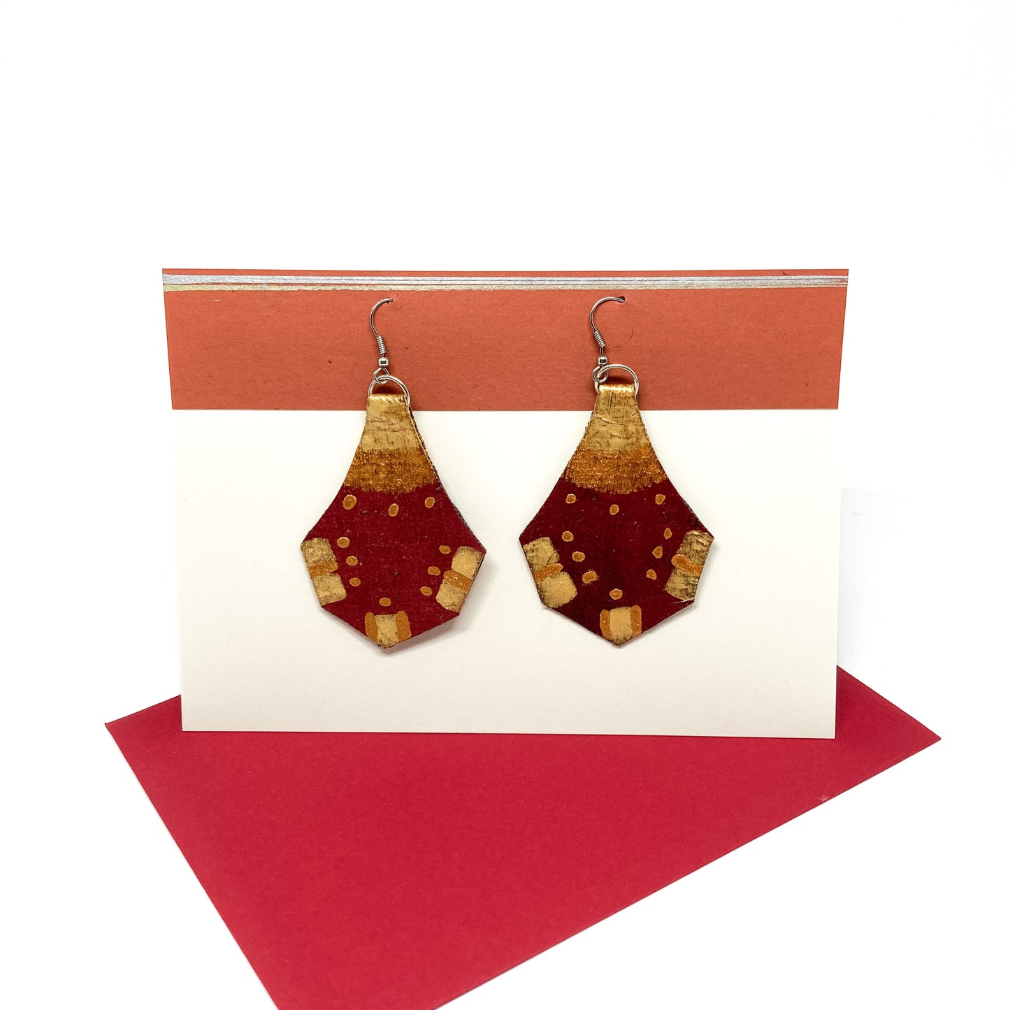 Two red and gold geometric earrings with a unique shape hang from silver hooks on a display card.