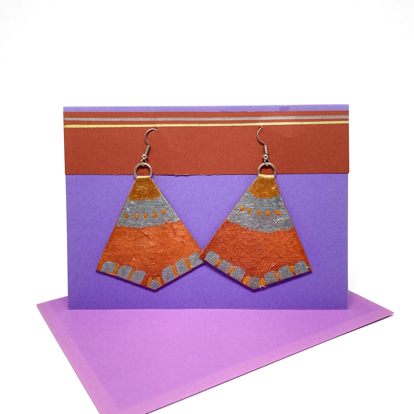 Two textured orange and silver geometric earrings hang from silver hooks on a display card with gold accents.