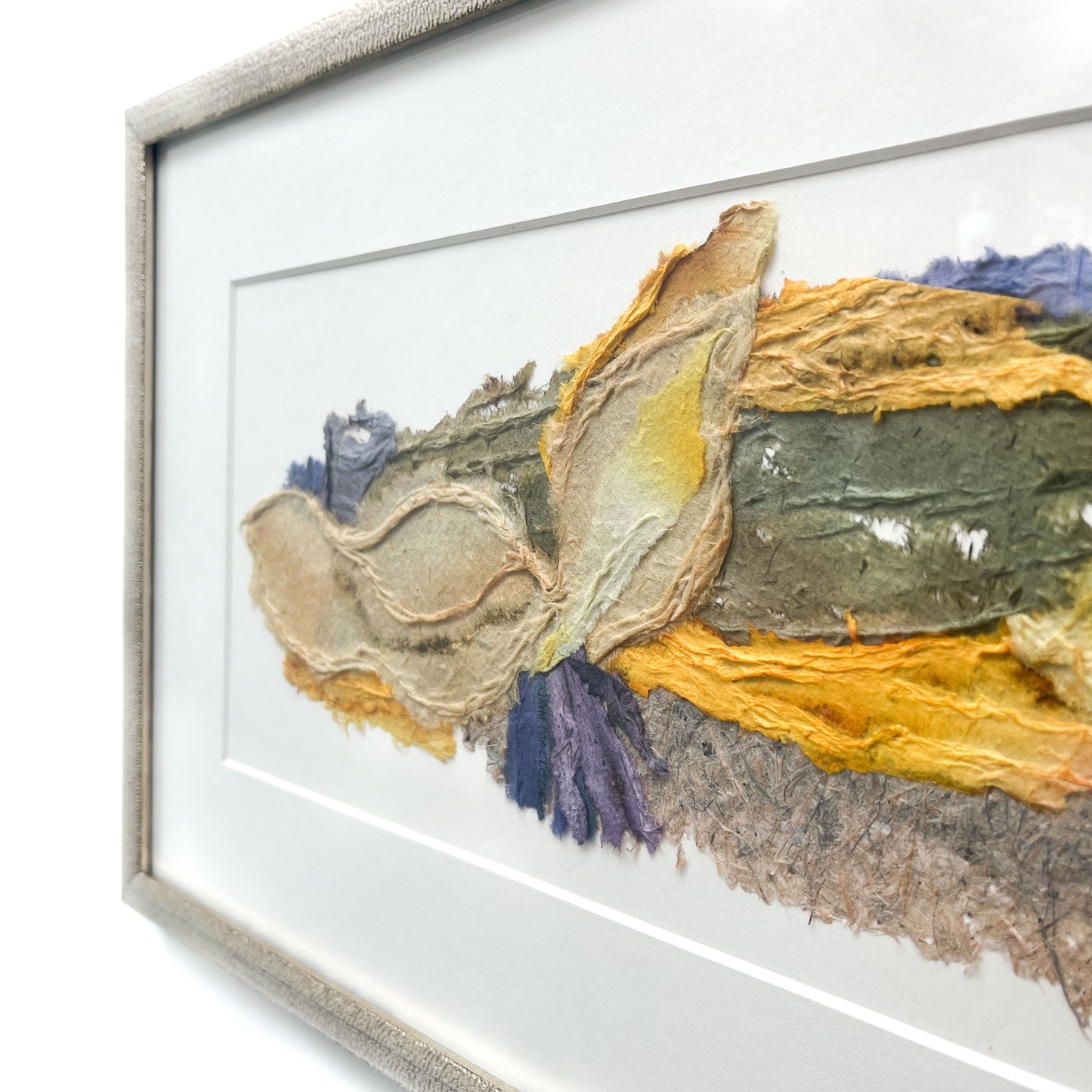 Angle view of framed horizontal abstract textile art with layered textures and blended hues of yellow, blue, green, brown, and gray.