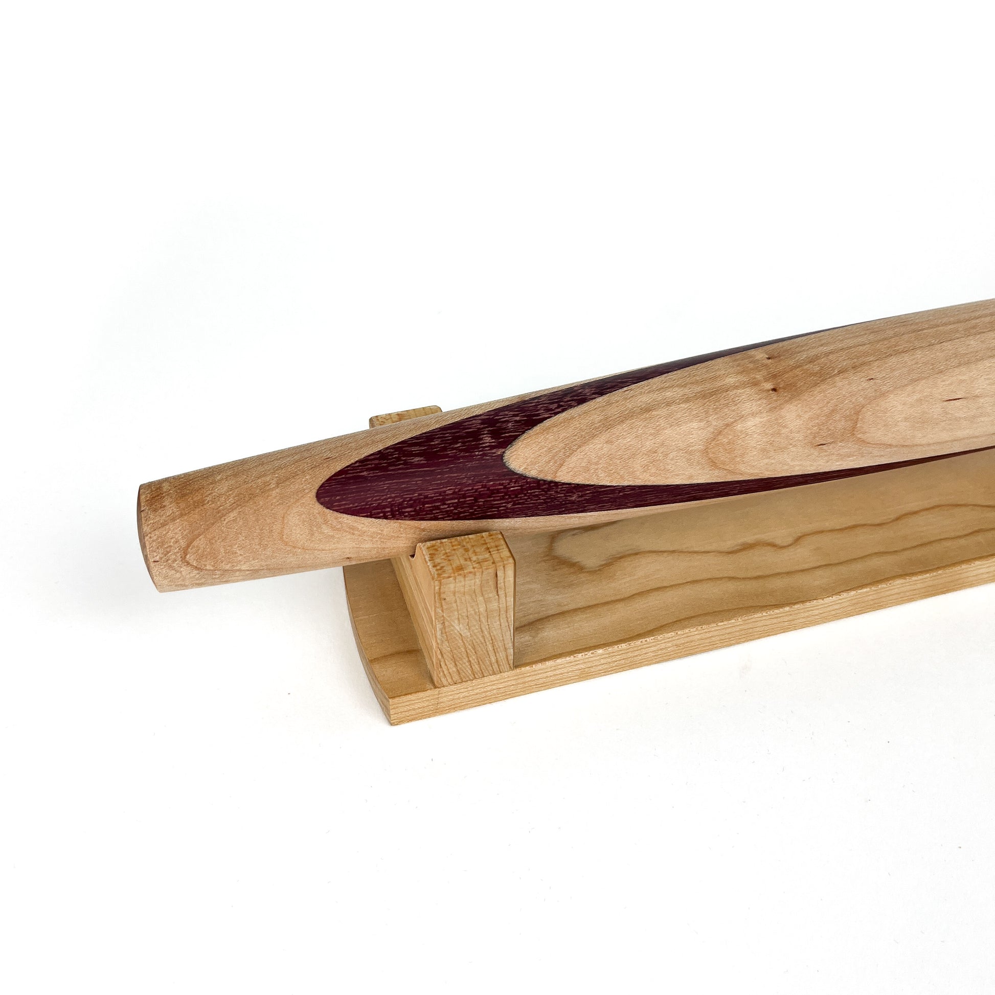 A close-up of a light-colored wooden French rolling pin with a dark inlay, resting on a wooden stand. The pin has a smooth, polished finish.