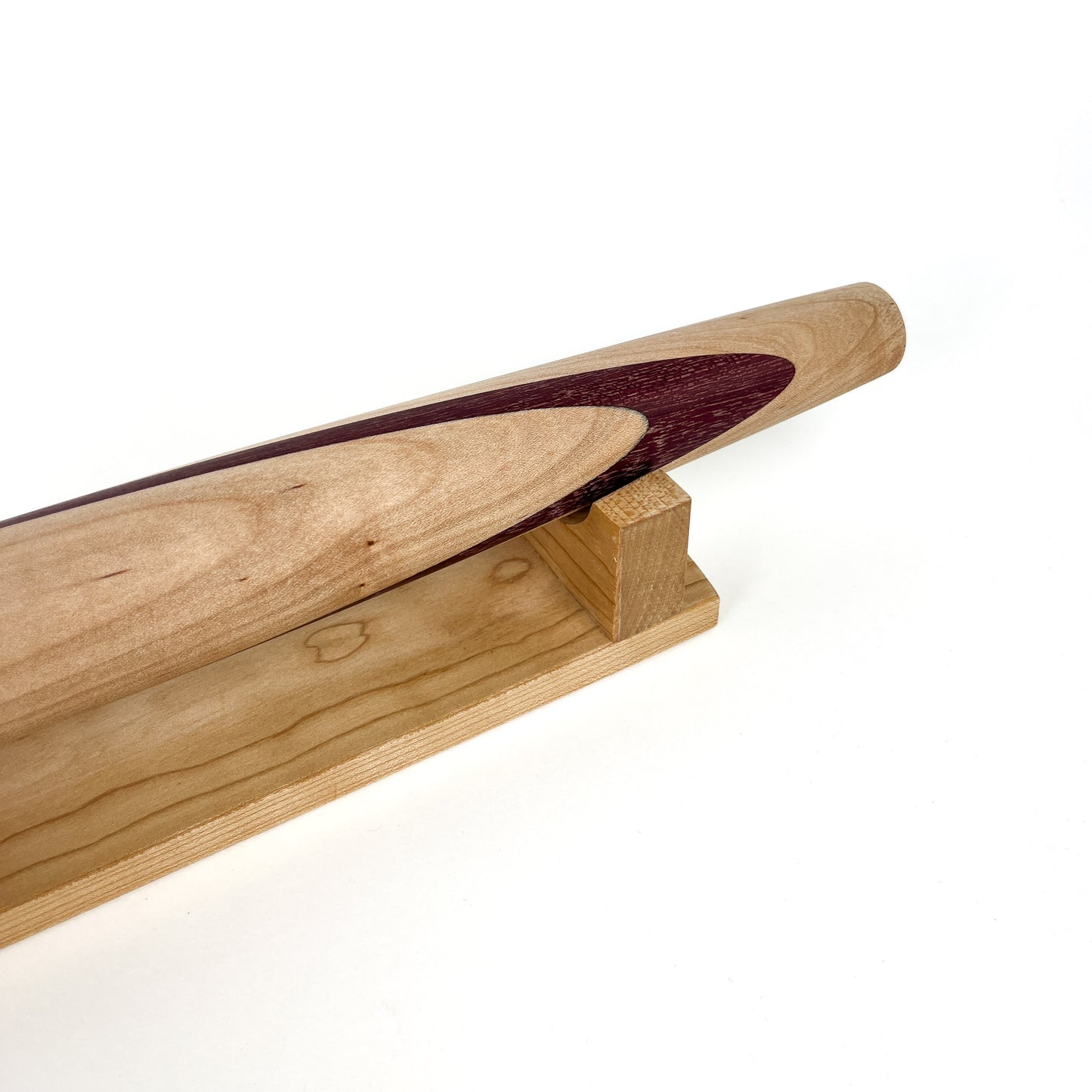 A close-up of a light-colored wooden French rolling pin with a dark inlay, resting on a wooden stand. The pin has a smooth, polished finish.