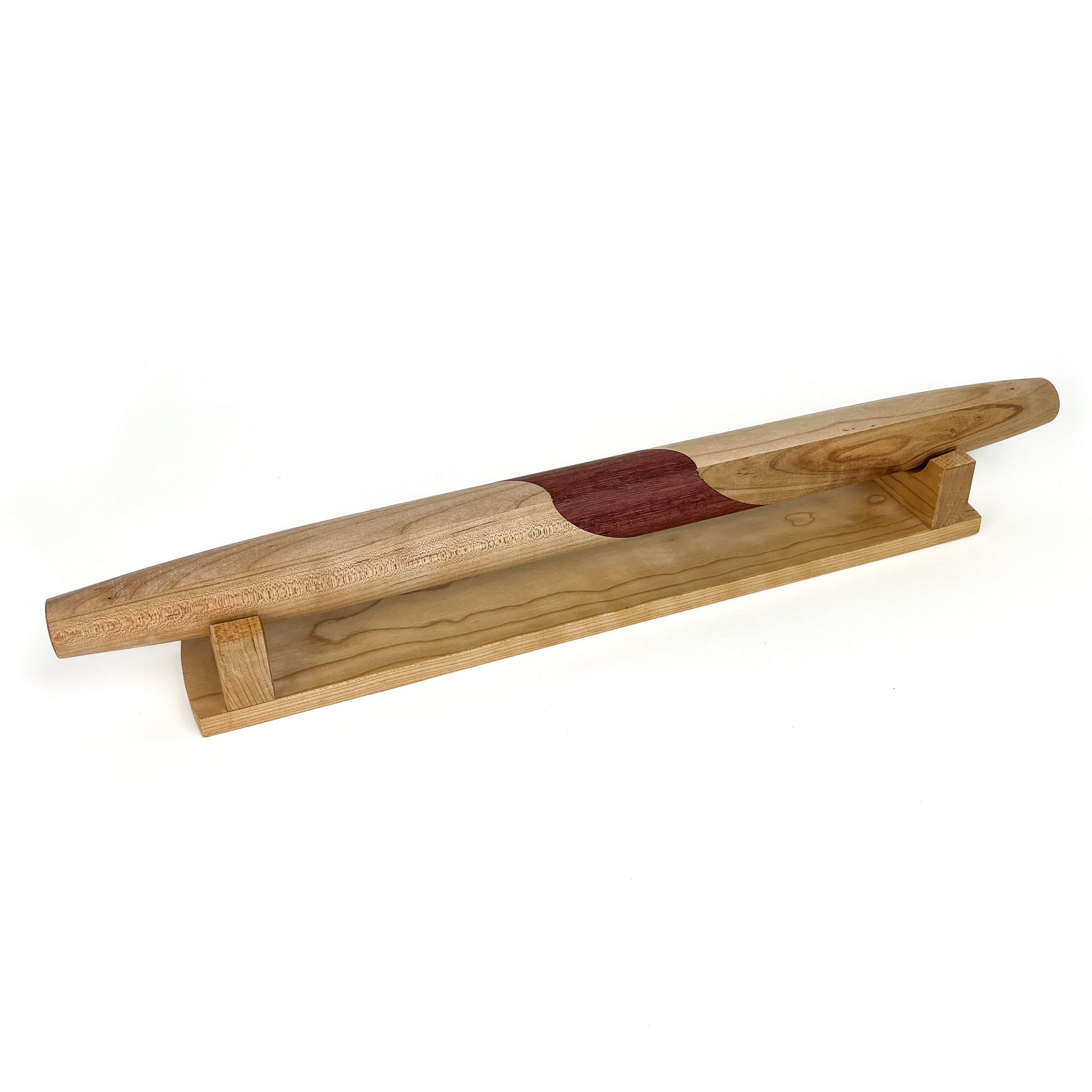 A wooden French rolling pin with a dark, curved inlay, resting horizontally on a light-colored wooden stand. The pin has a smooth finish.