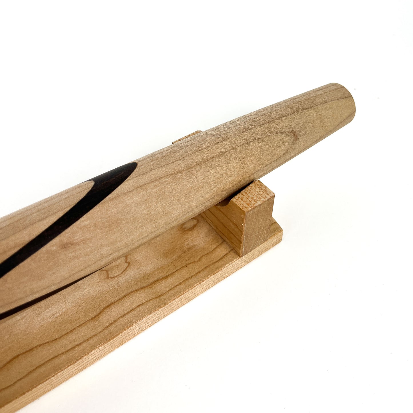 A close-up of a Sugar Maple French rolling pin with a Black Walnut Celtic knot inlay, resting on a wooden stand. The pin has a smooth finish.