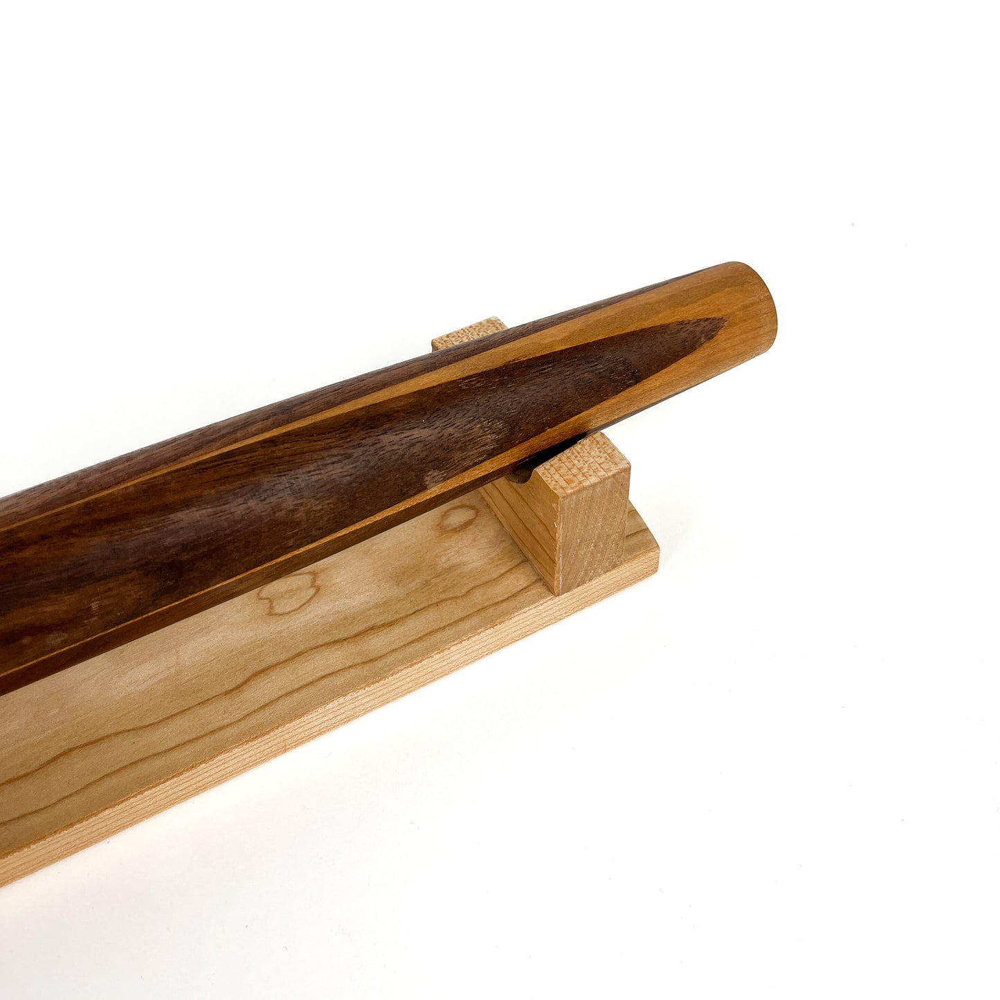 A close-up of a French rolling pin made of Black Walnut wood, resting on a Sugar Maple stand. The pin has a smooth, polished finish.